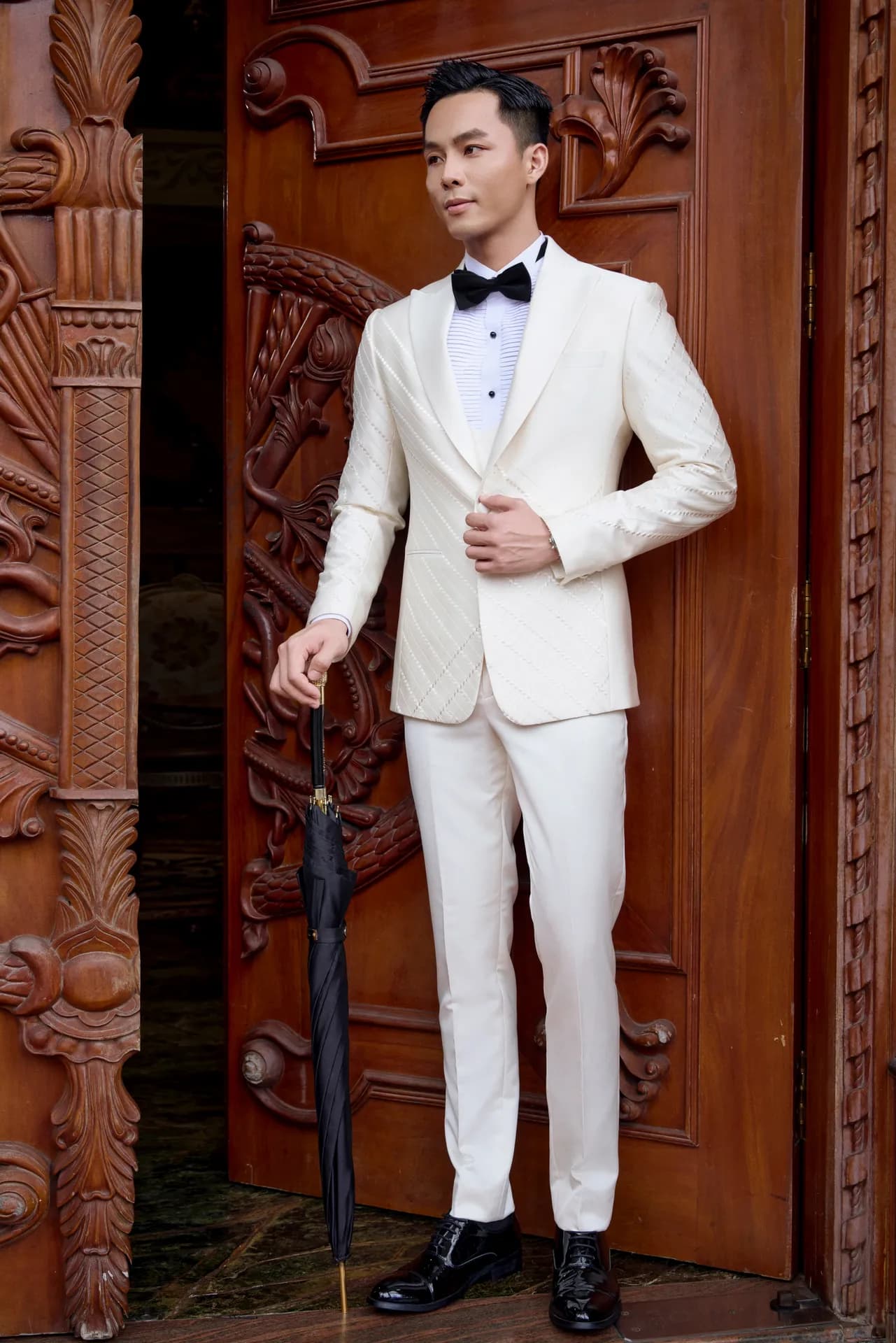 Tuxedo trắng kem, cầm ô đen, đứng bên cửa gỗ chạm khắc