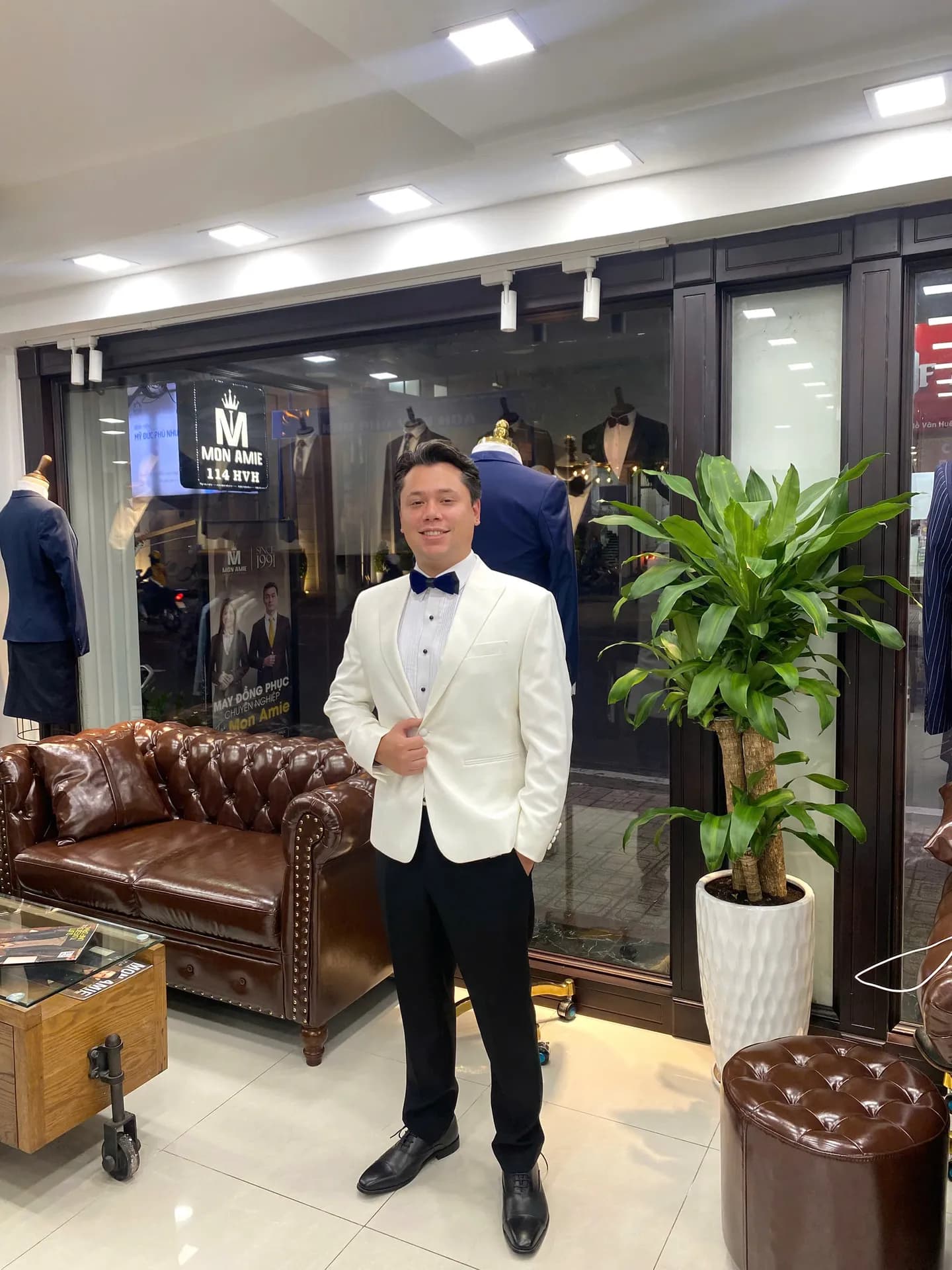 Khách mặc dinner jacket trắng, nơ xanh navy, cạnh sofa da showroom