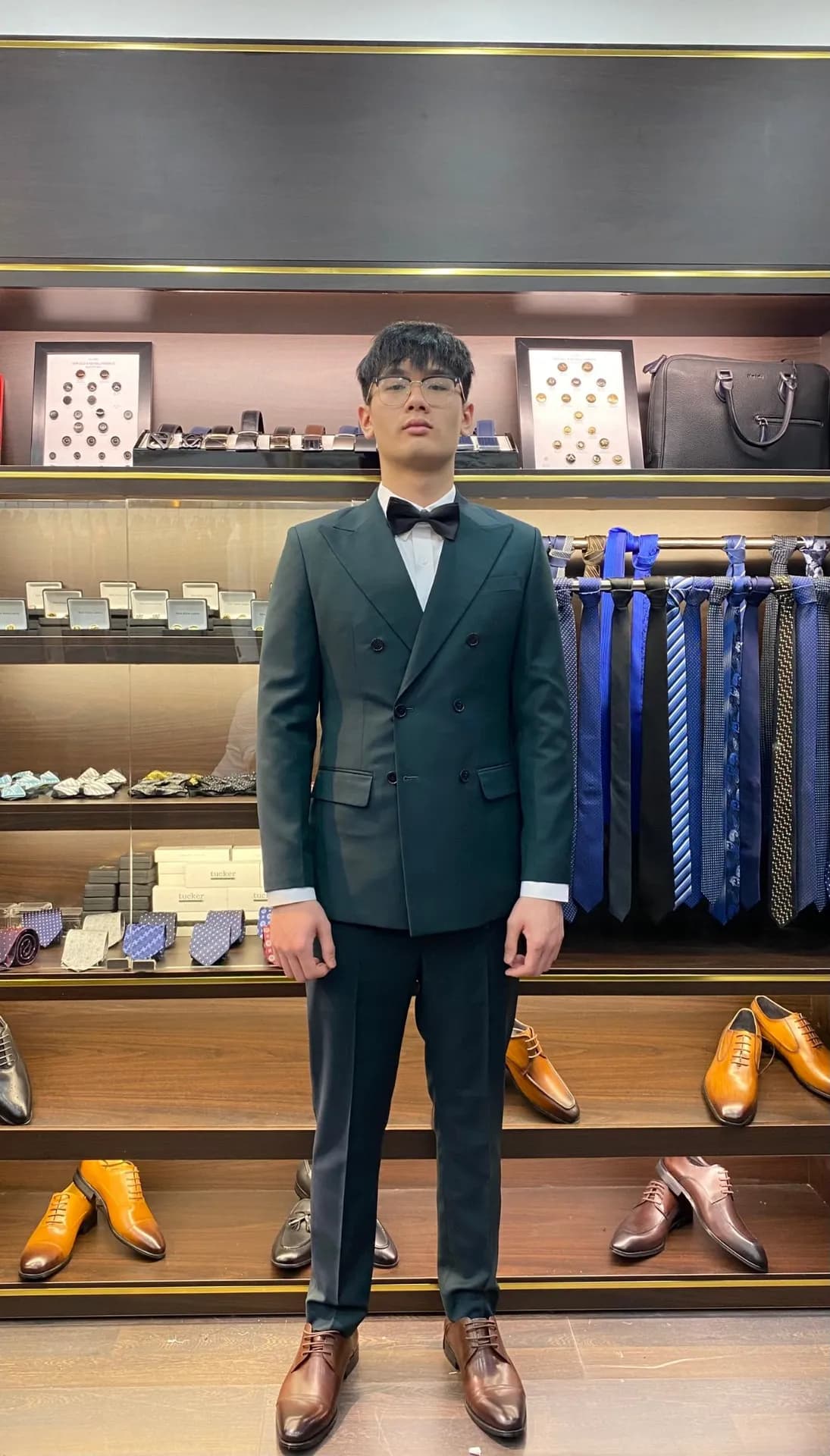 Khách mặc suit xanh rêu cài hai hàng, nơ đen, trước kệ phụ kiện