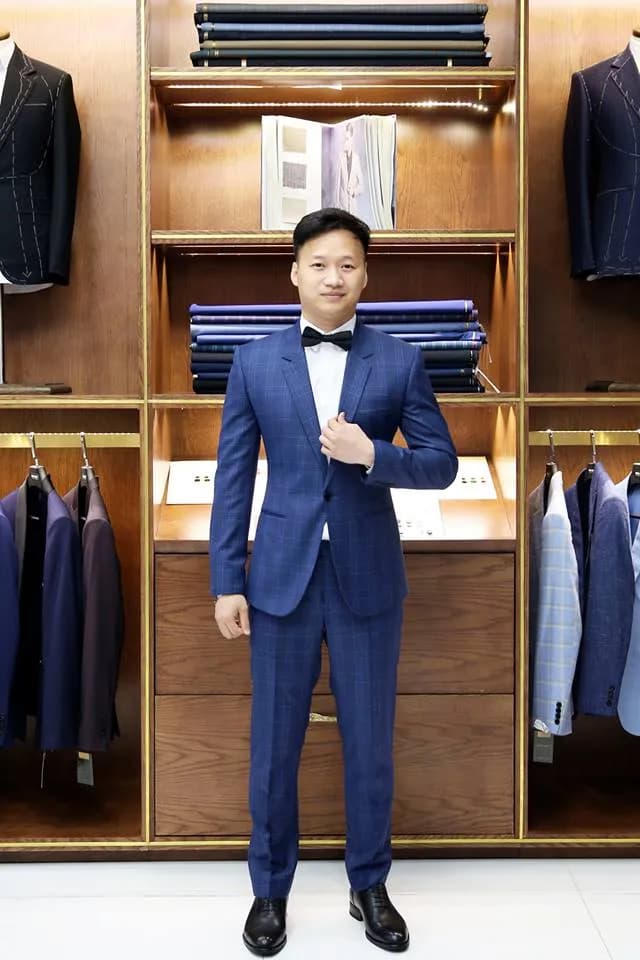 Khách mặc suit xanh ca-rô chìm, nơ đen, chỉnh khuy trước tủ vải