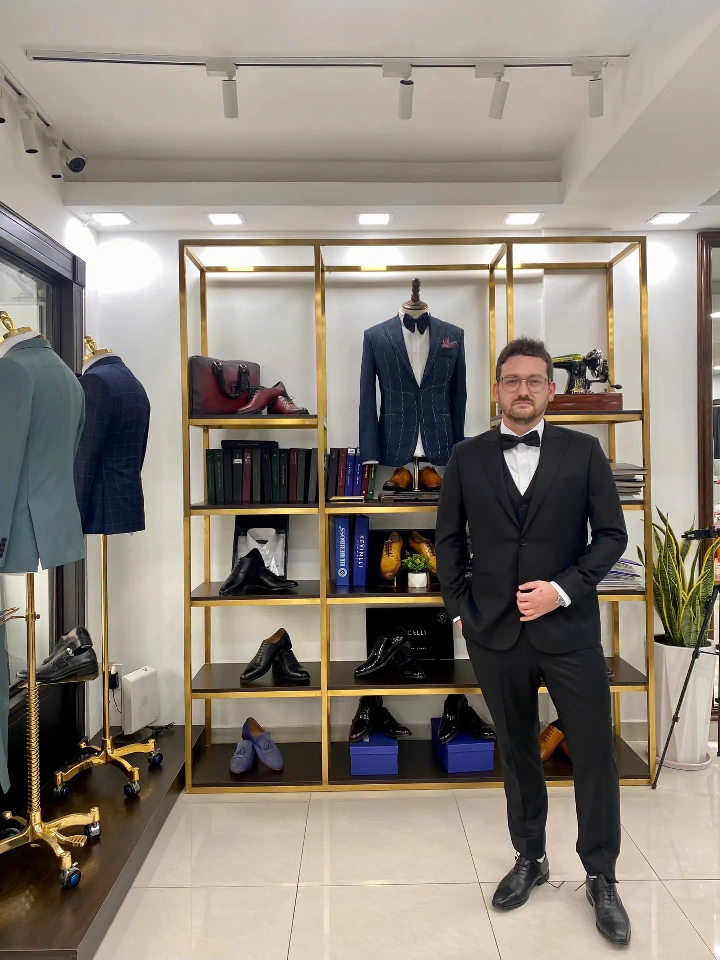 Khách mặc tuxedo ba mảnh nơ đen, đứng cạnh kệ phụ kiện vàng đồng