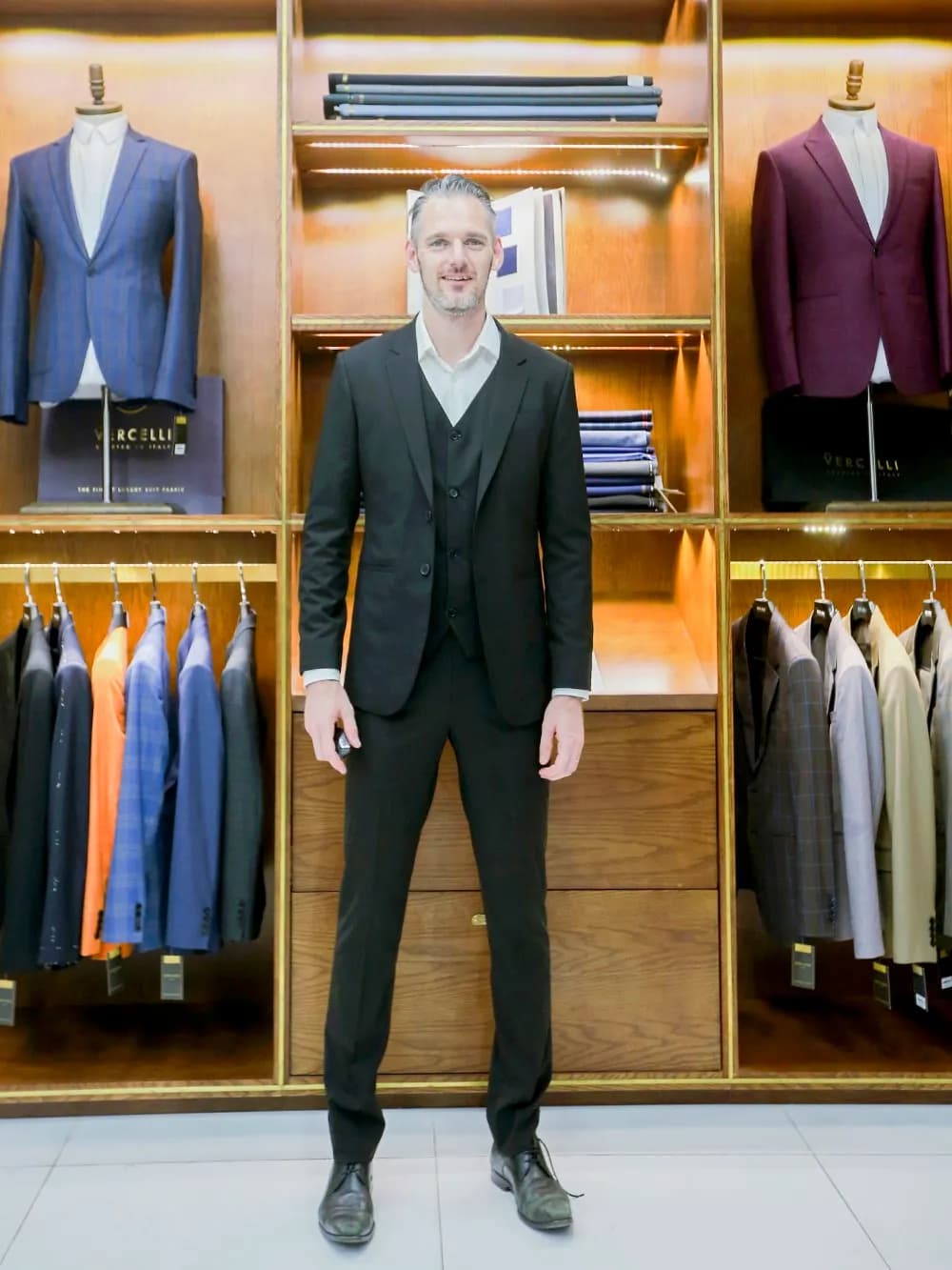 Khách mặc suit ba mảnh xanh rêu đậm trước tủ trưng bày vest