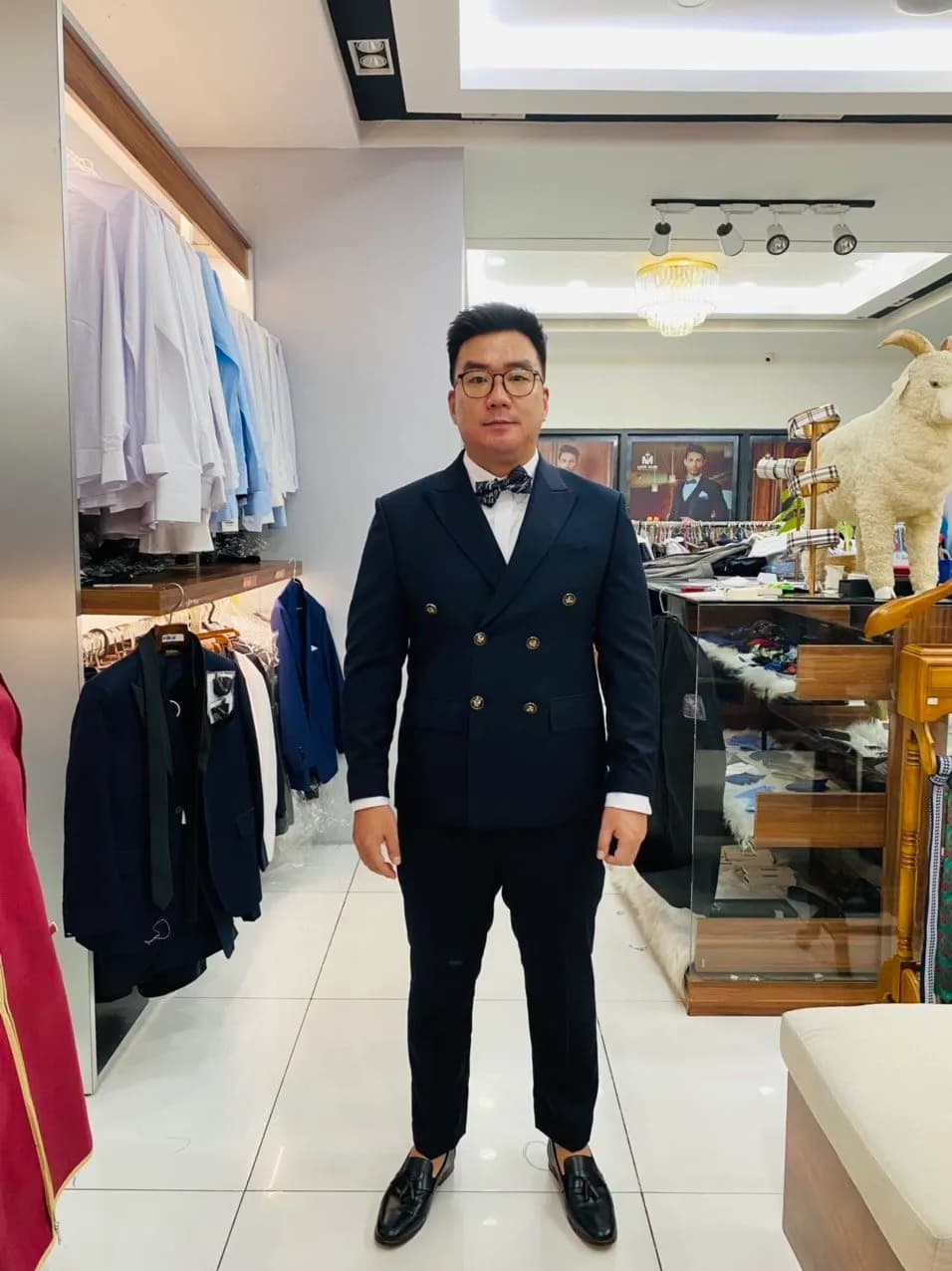 Khách mặc vest hai hàng navy, nơ đen, mỉm cười tại showroom