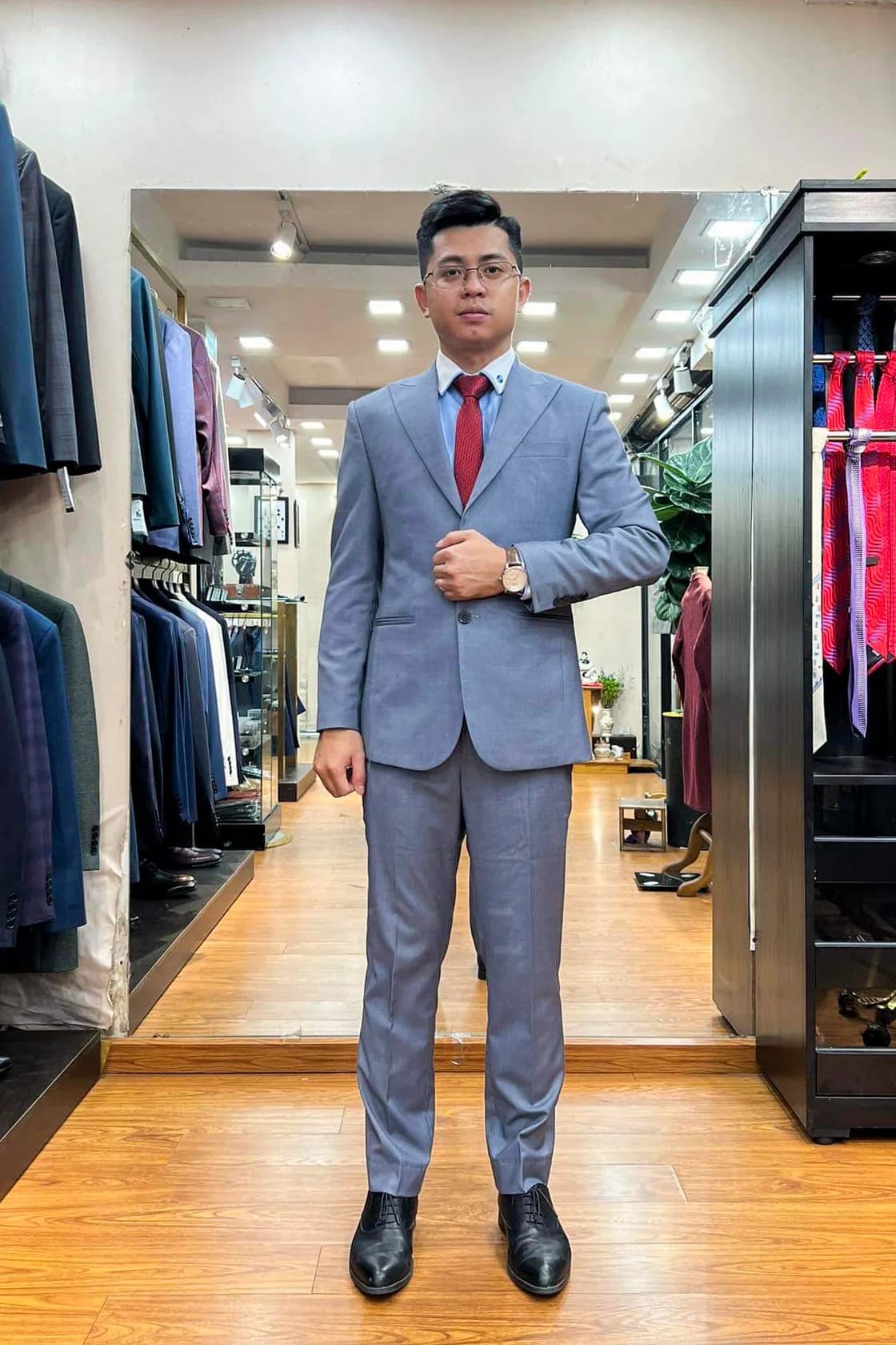 Khách mặc suit xanh xám nhạt, cà vạt đỏ, chỉnh khuy tay áo