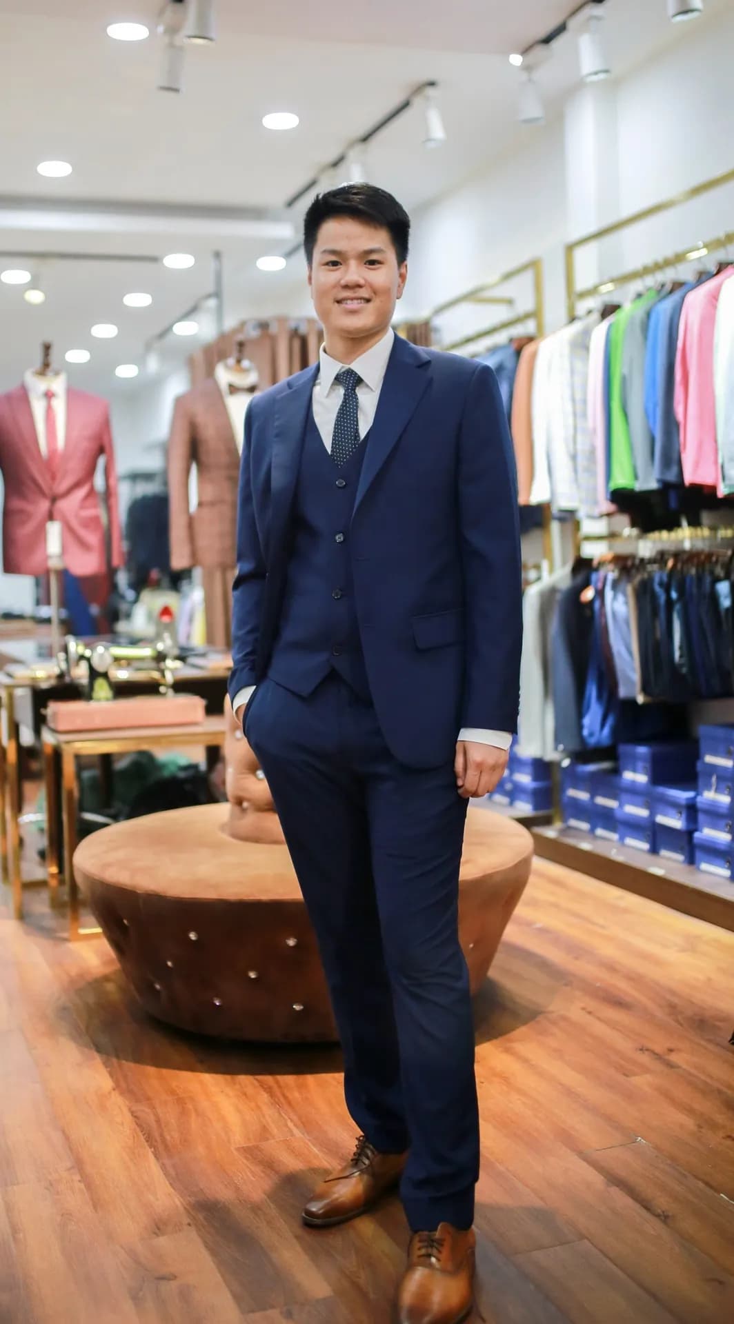 Khách mặc suit ba mảnh navy, tay đút túi, showroom sàn gỗ