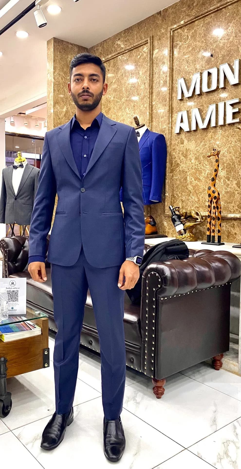 Khách mặc suit xanh dương sơ mi đen, đứng cạnh sofa trước biển Mon Amie
