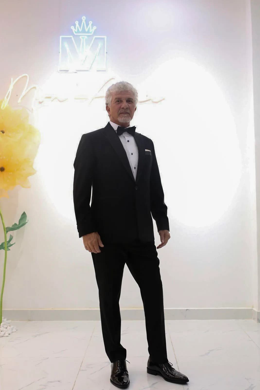 Khách lớn tuổi mặc tuxedo nơ đen trước tường logo Mon Amie sáng