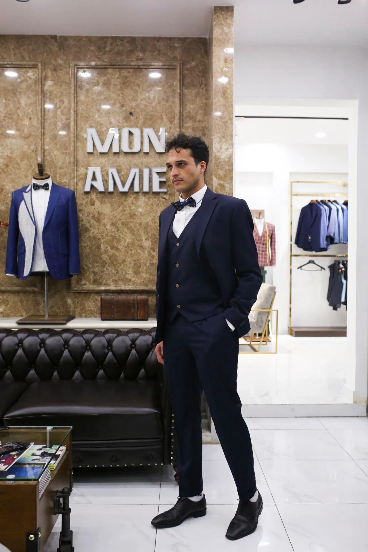 Khách mặc suit ba mảnh navy, nơ đen, bên cạnh mannequin tuxedo xanh