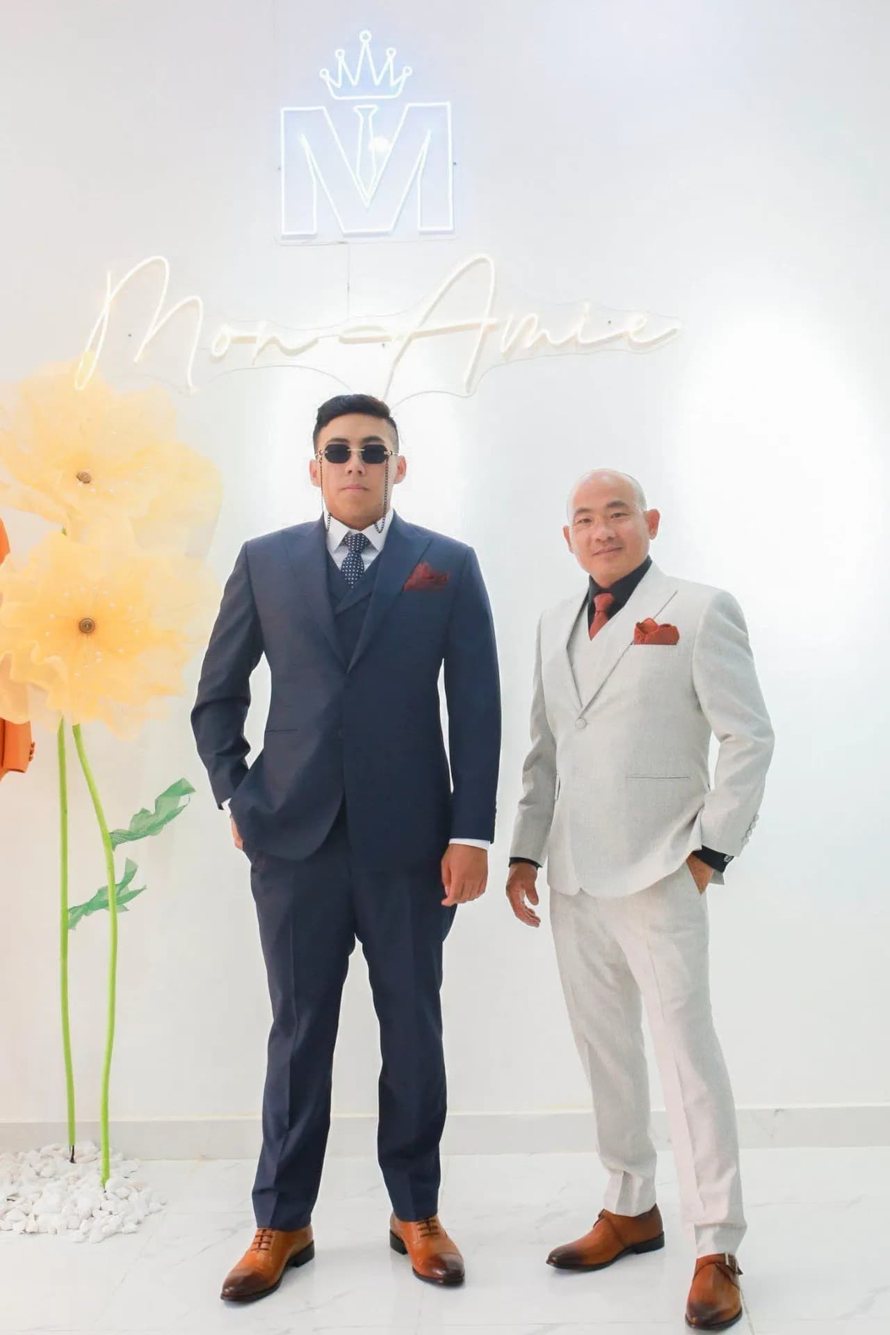 Hai khách mặc suit navy và suit trắng trước tường logo Mon Amie