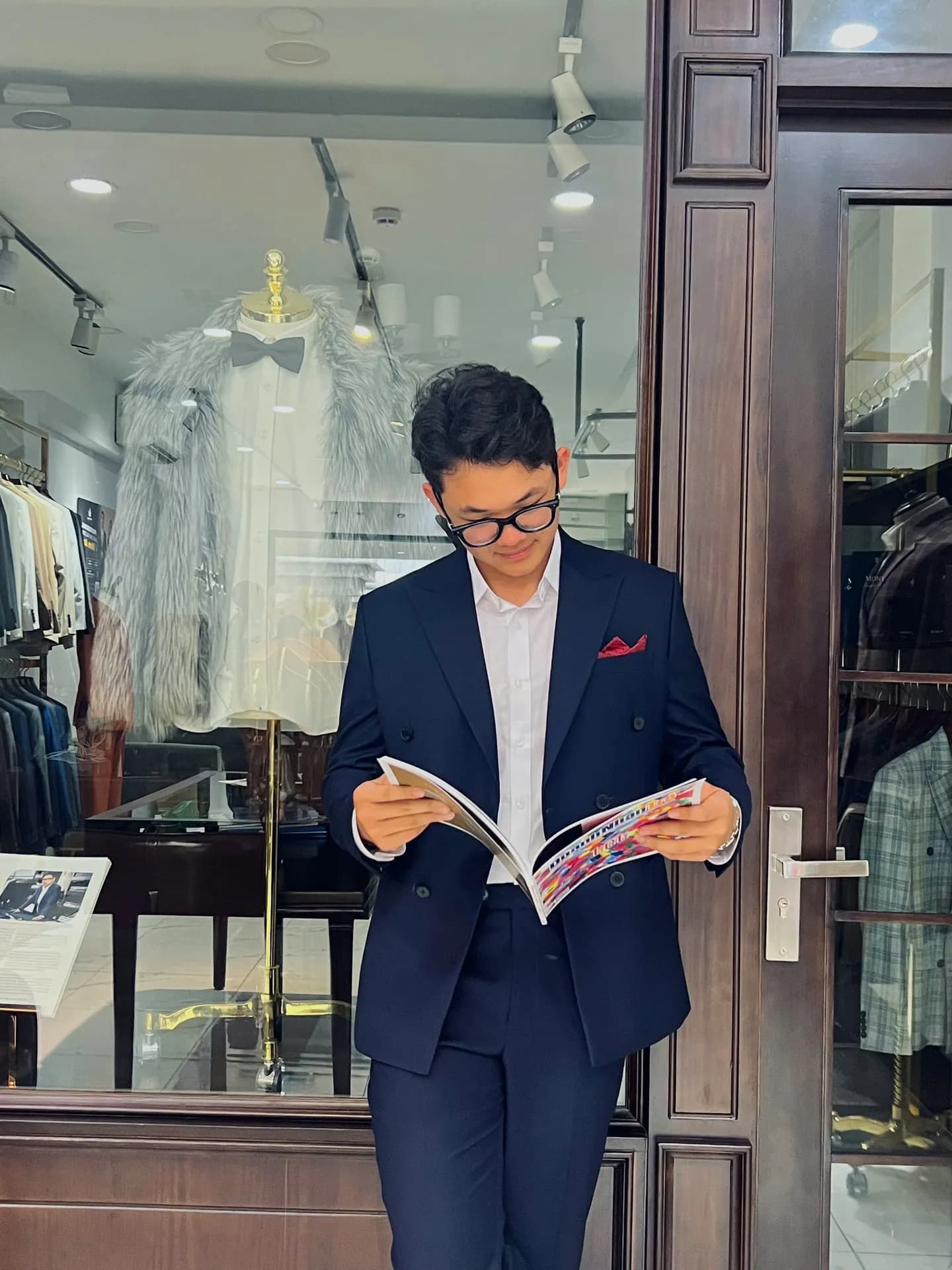 Khách mặc vest navy hai hàng, khăn túi đỏ, lật xem tạp chí