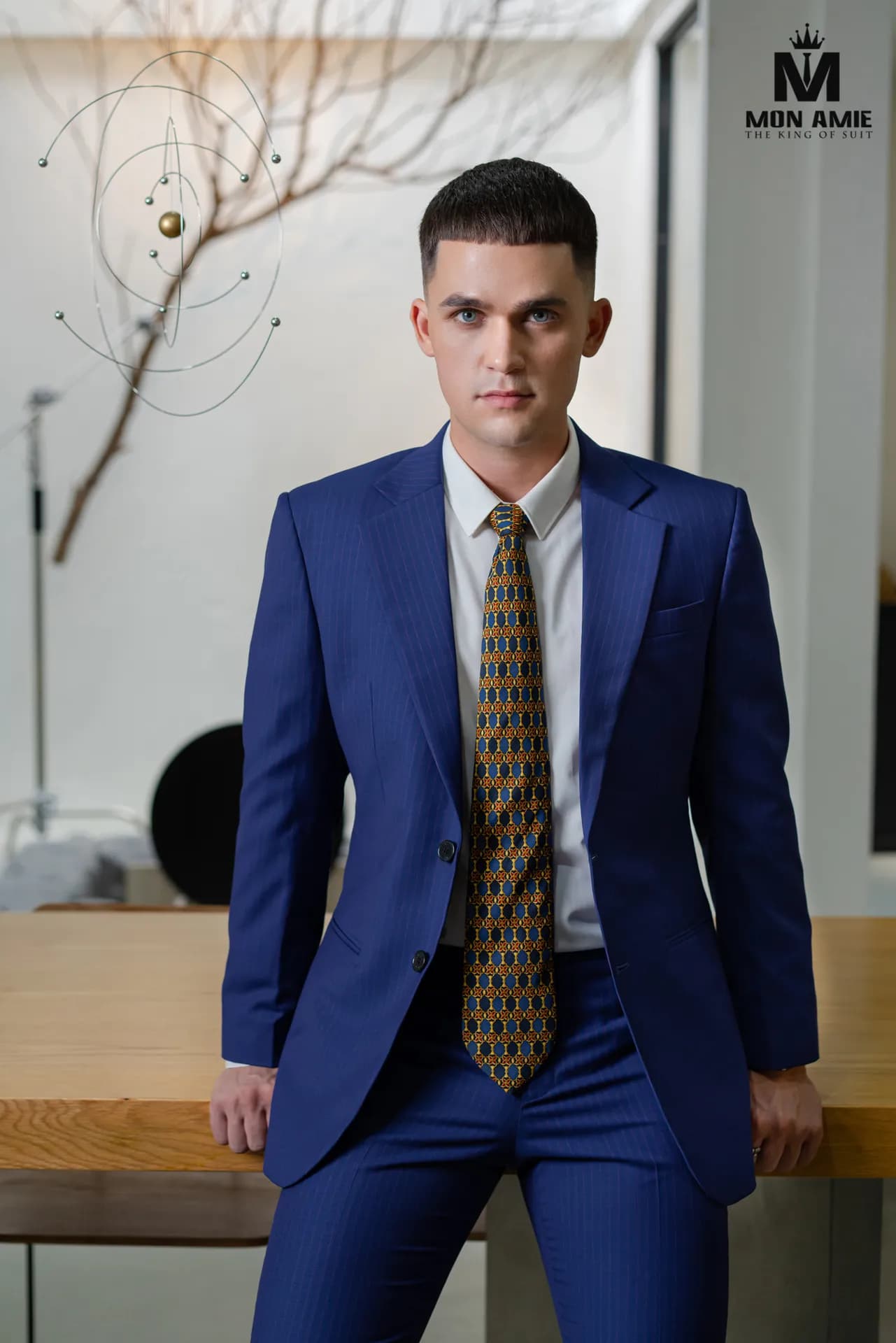 Suit xanh sapphire kẻ sọc mảnh, cà vạt họa tiết vàng, dựa bàn gỗ