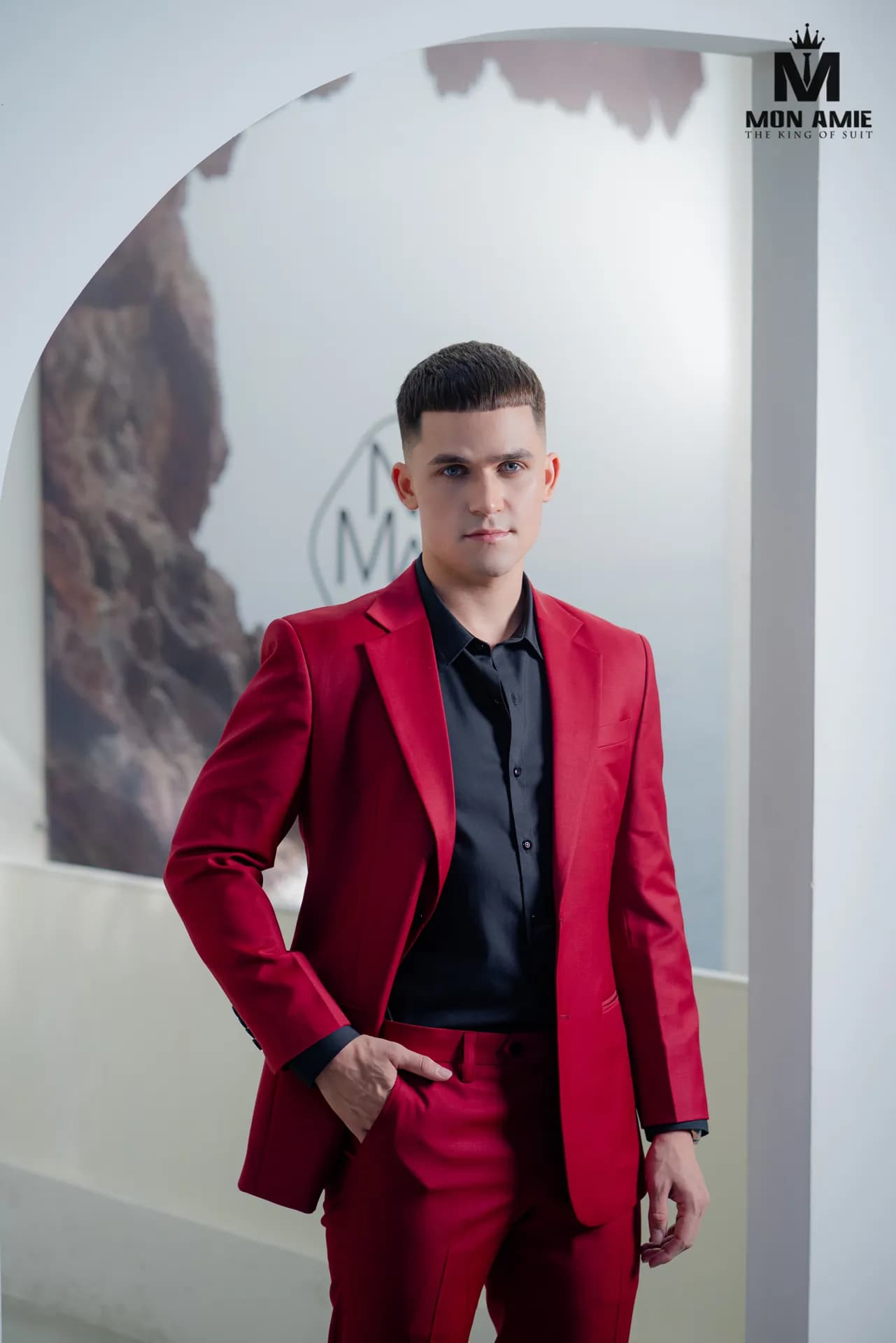 Suit đỏ đô, sơ mi đen, tay đút túi quần trước khung vòm trắng