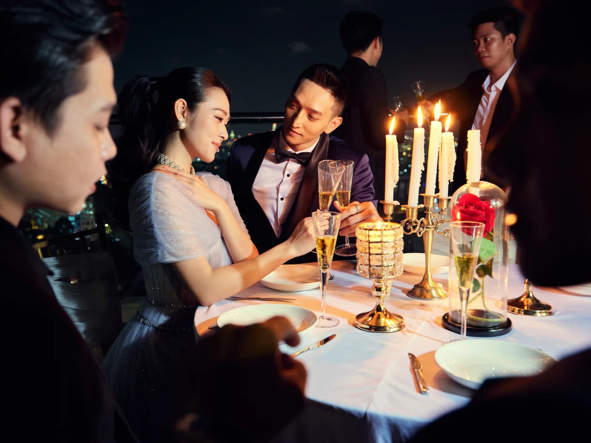Bữa tối lãng mạn trên sân thượng: tuxedo navy, nến và hoa hồng bên ly champagne