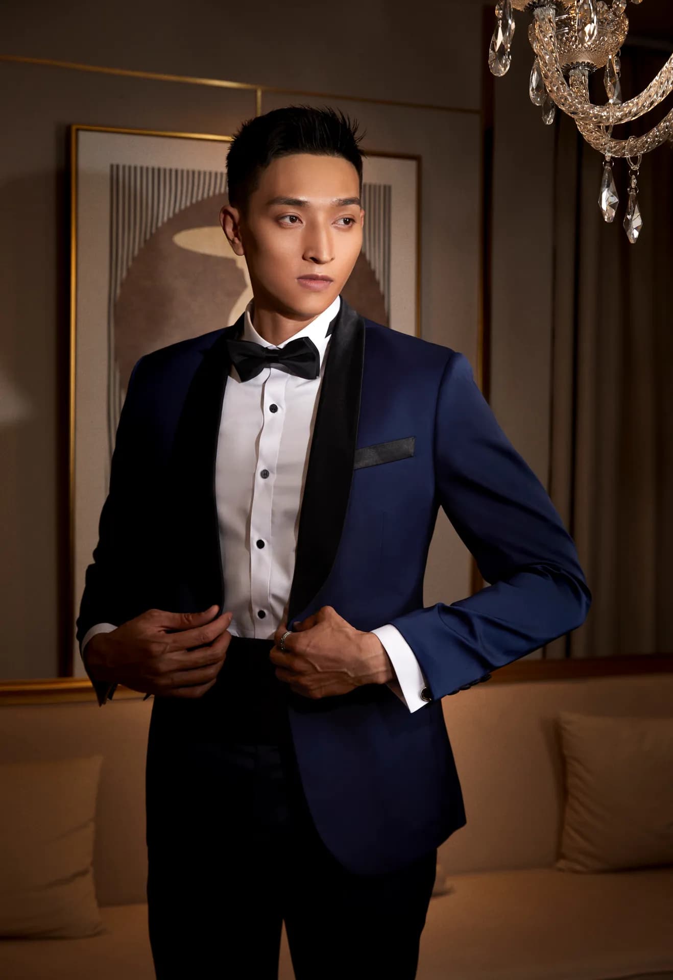 Tuxedo xanh sapphire ve nhung đen, hai tay chỉnh vạt áo, ánh nhìn nghiêng