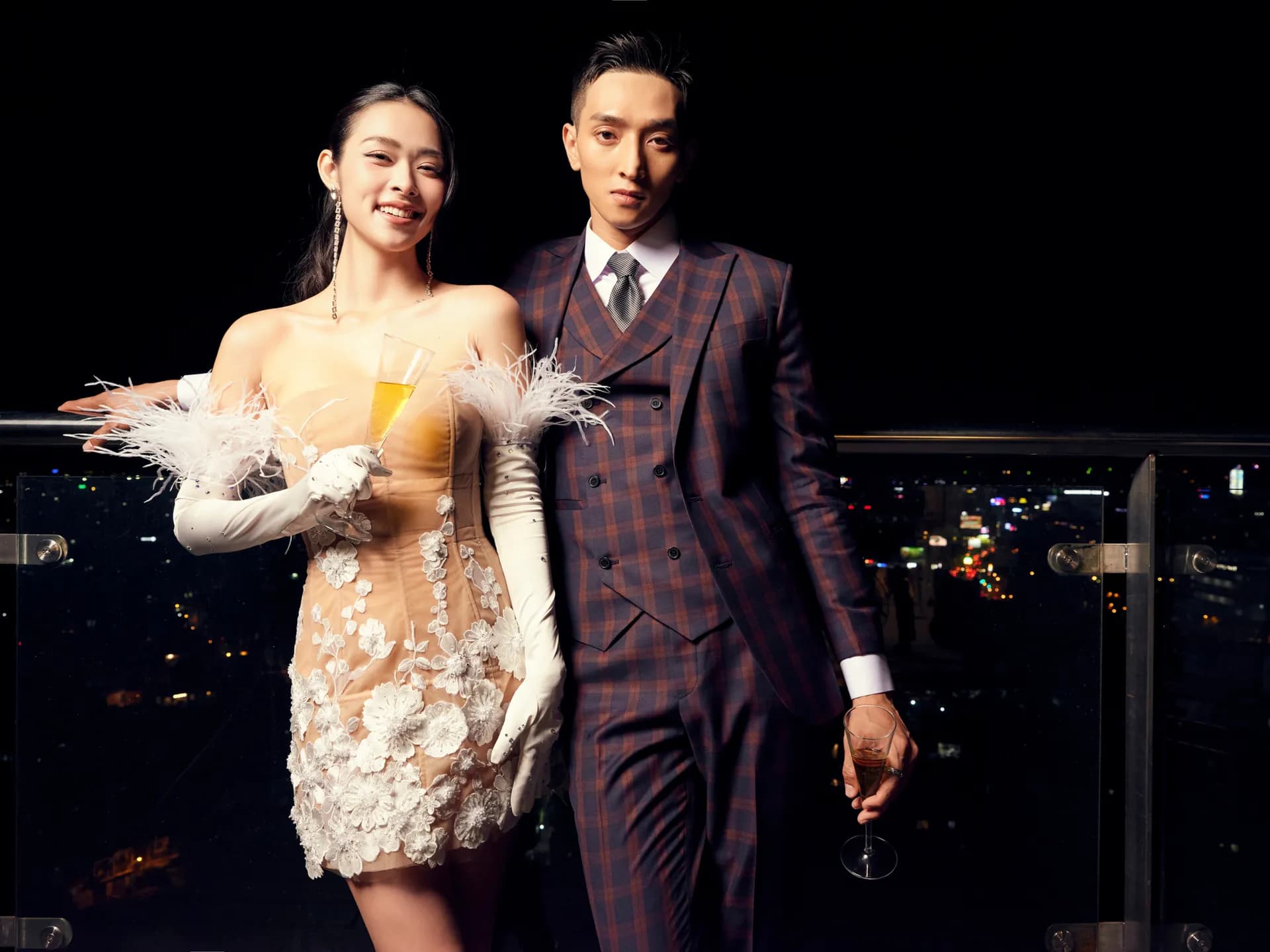 Suit ba mảnh kẻ ô tím cầm ly, sánh đôi váy dạ tiệc trên sân thượng đêm