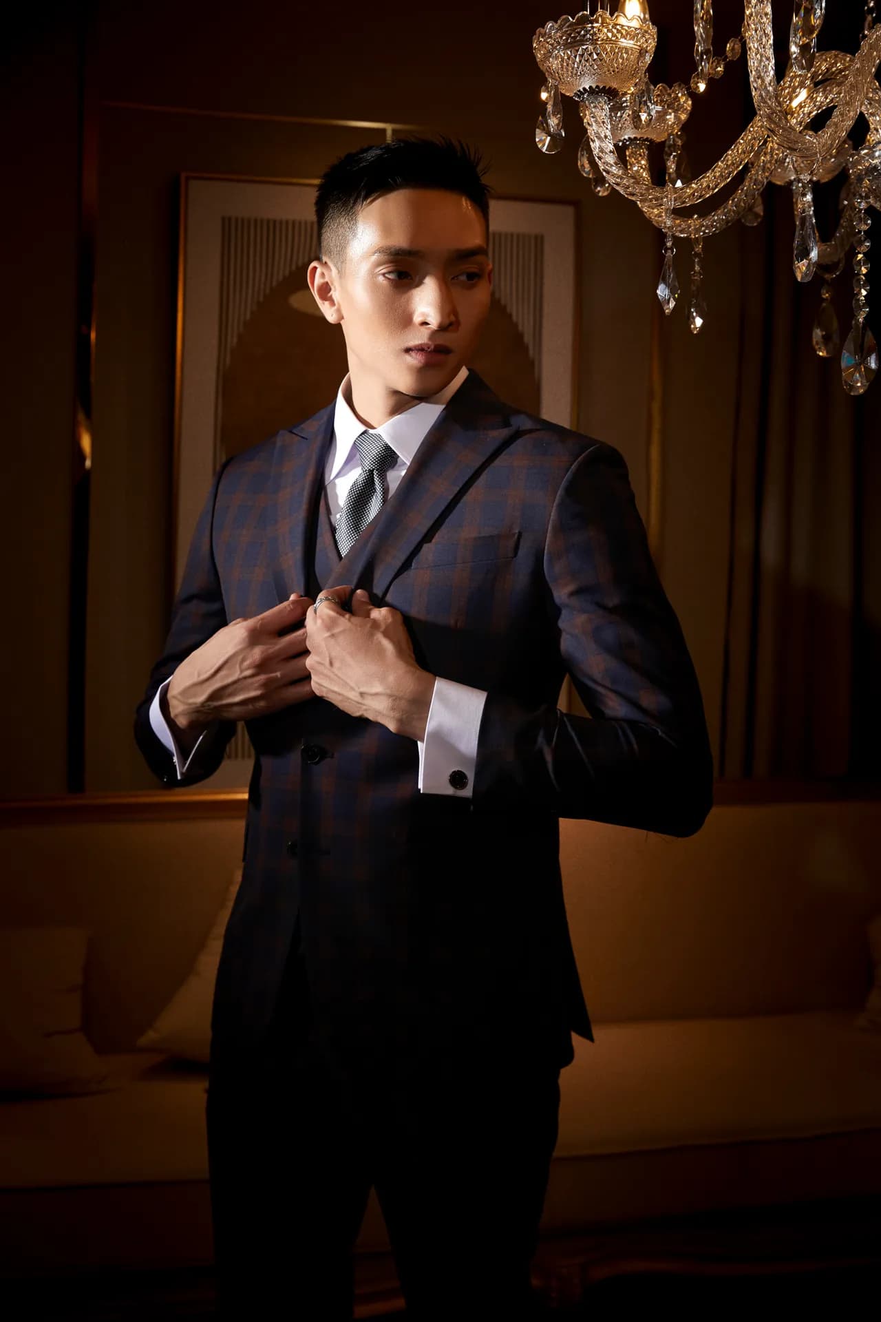 Suit ba mảnh kẻ ô tối màu, hai tay chỉnh vạt áo, nhìn nghiêng