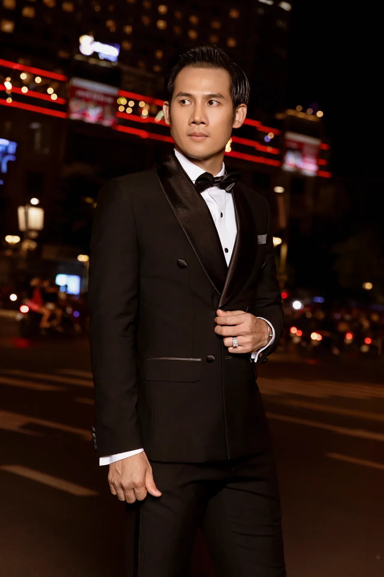 Tuxedo đen hai hàng khuy ve satin, chỉnh áo, nhìn nghiêng giữa phố đêm