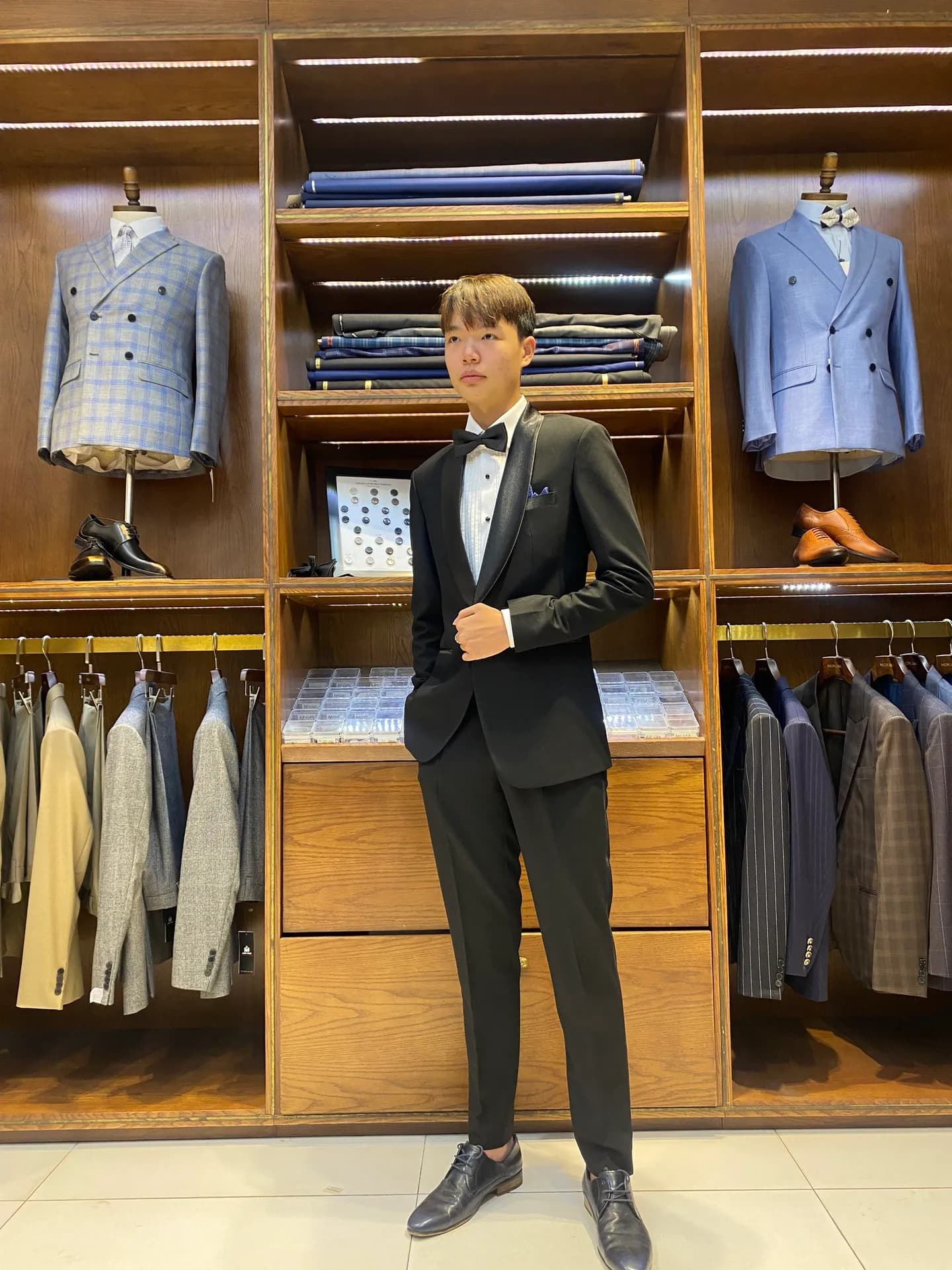 Tuxedo đen cổ lụa nơ đen, tay chỉnh áo trước kệ gỗ ấm cúng