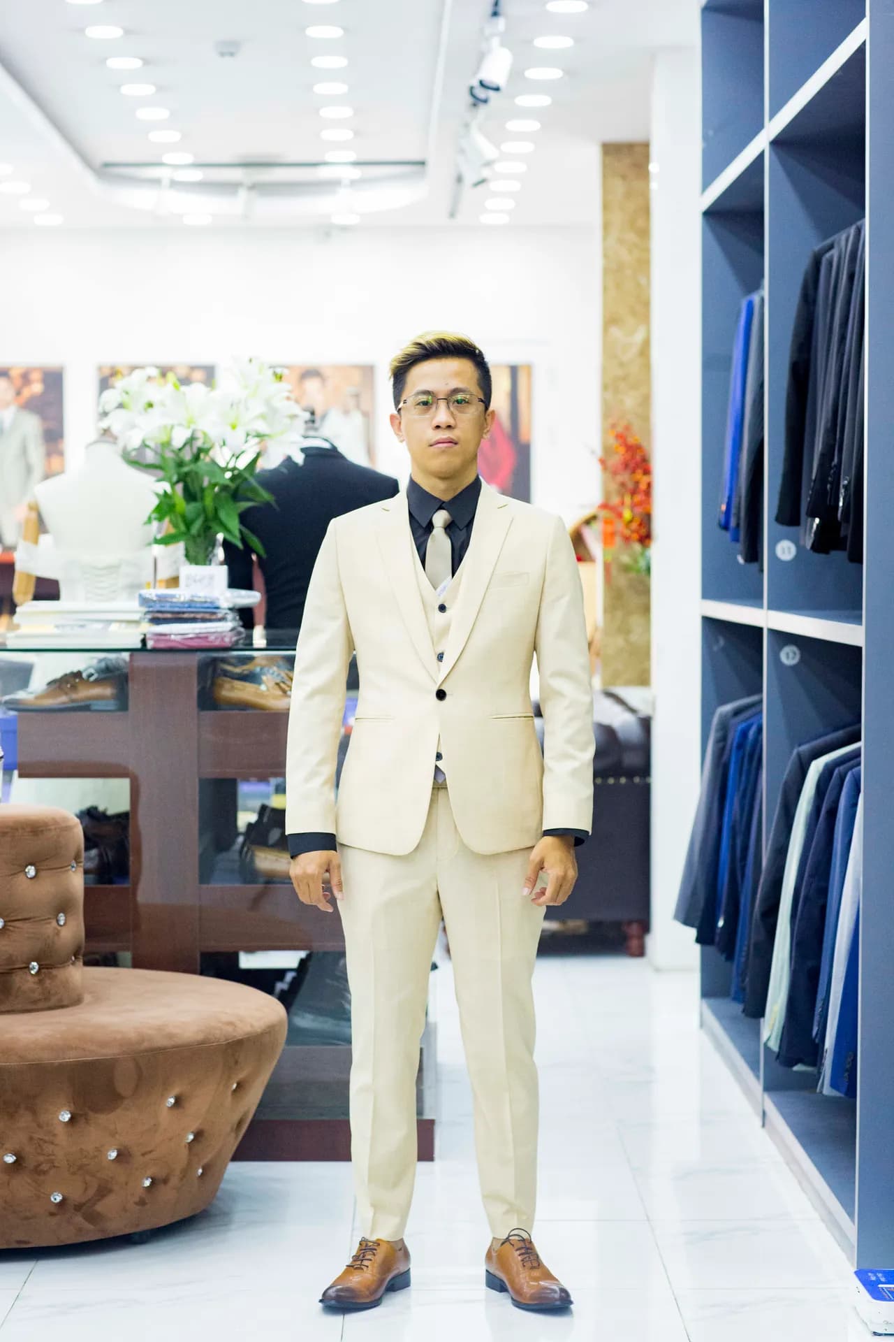 Khách mặc suit ba mảnh kem, sơ mi đen, đứng giữa showroom