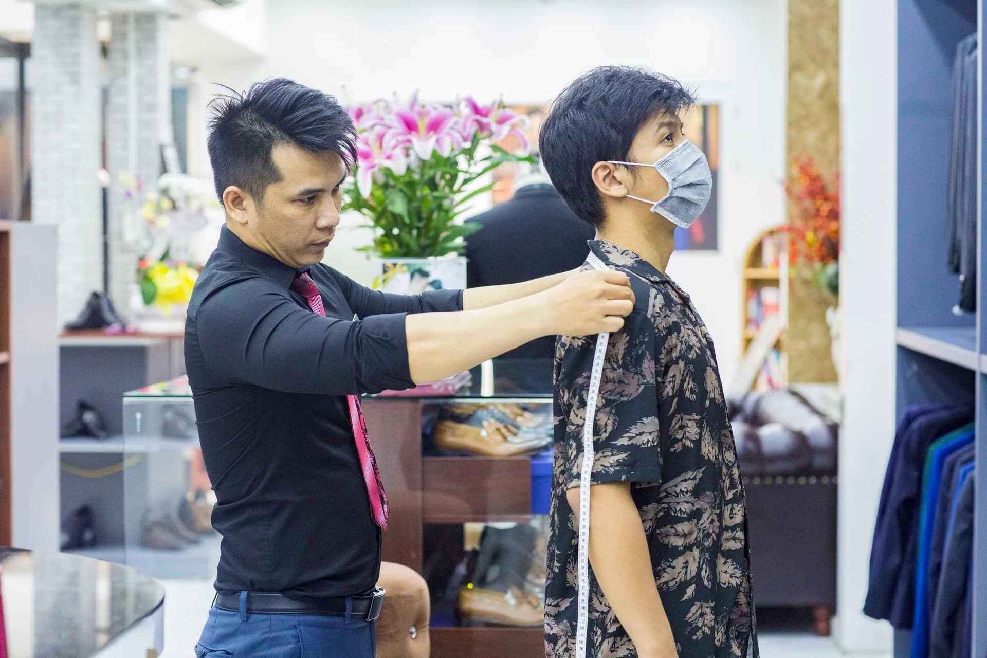Thợ may đo vai cho khách, nền hoa lan và kệ giày trong showroom