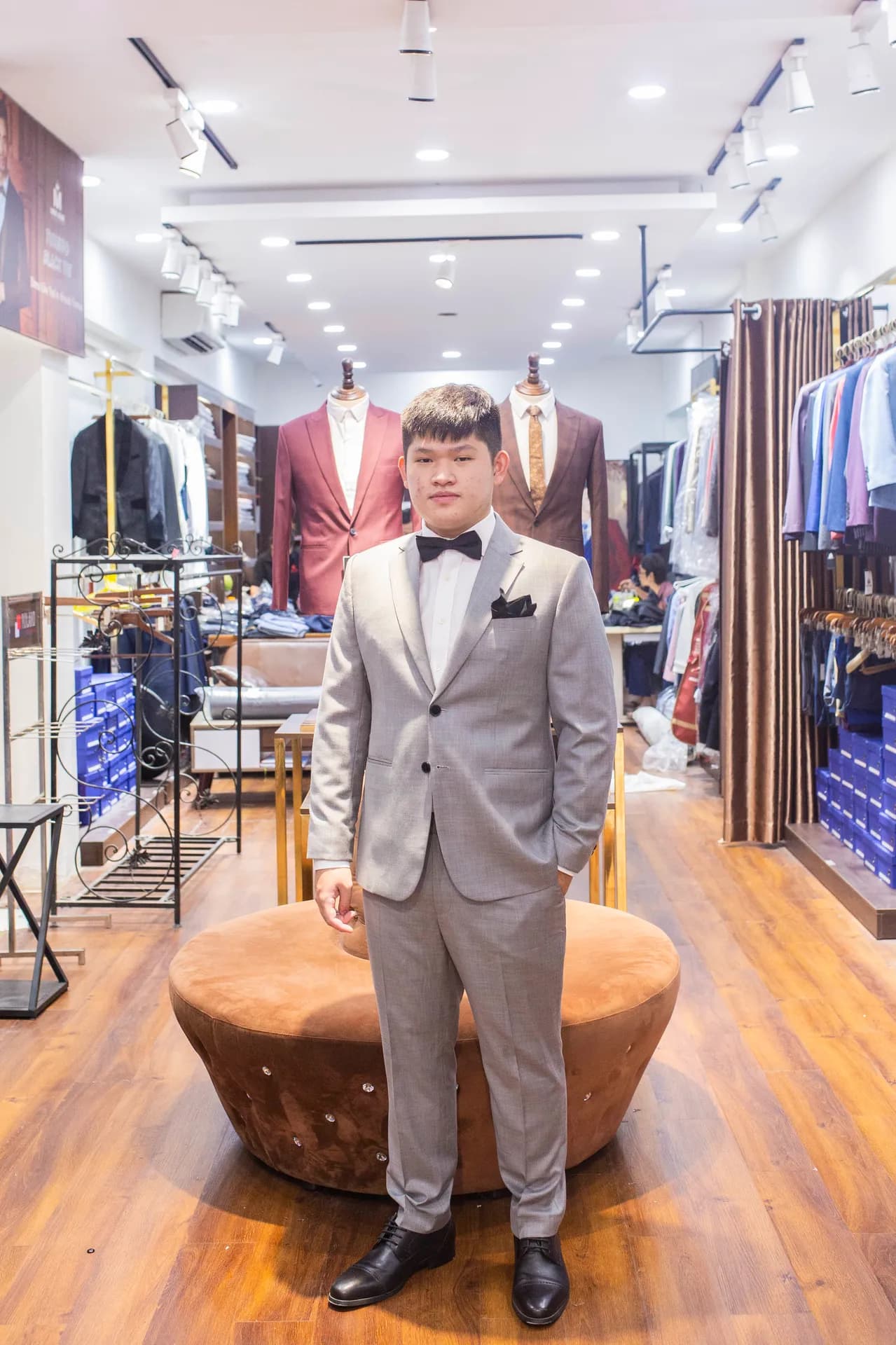 Khách mặc suit xám nhạt, nơ đen, khăn túi, đứng trên ghế nhung nâu