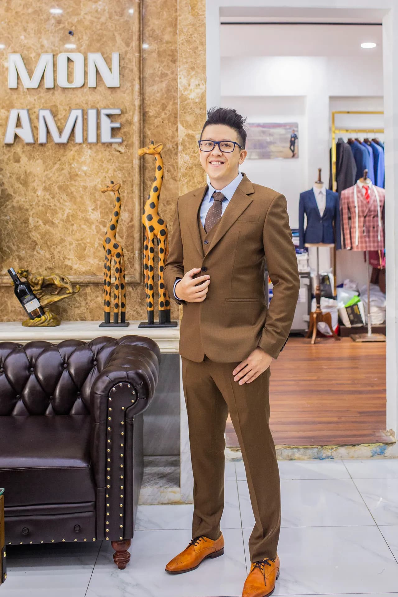 Suit nâu ba mảnh, giày da cam, đứng bên chữ MON AMIE và sofa Chesterfield