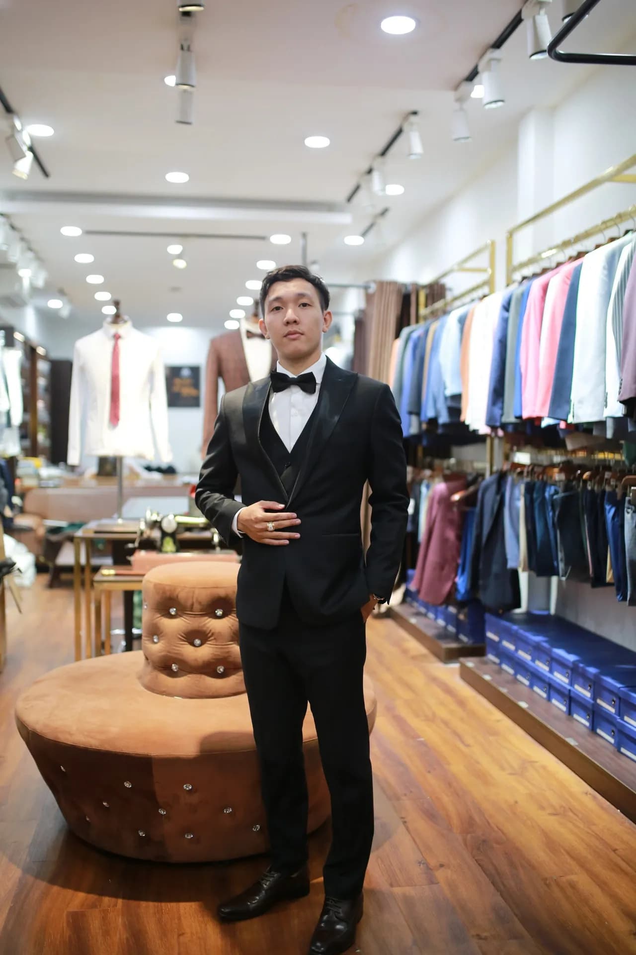 Tuxedo đen cổ nơ, ve áo satin, dáng đứng giữa showroom ấm