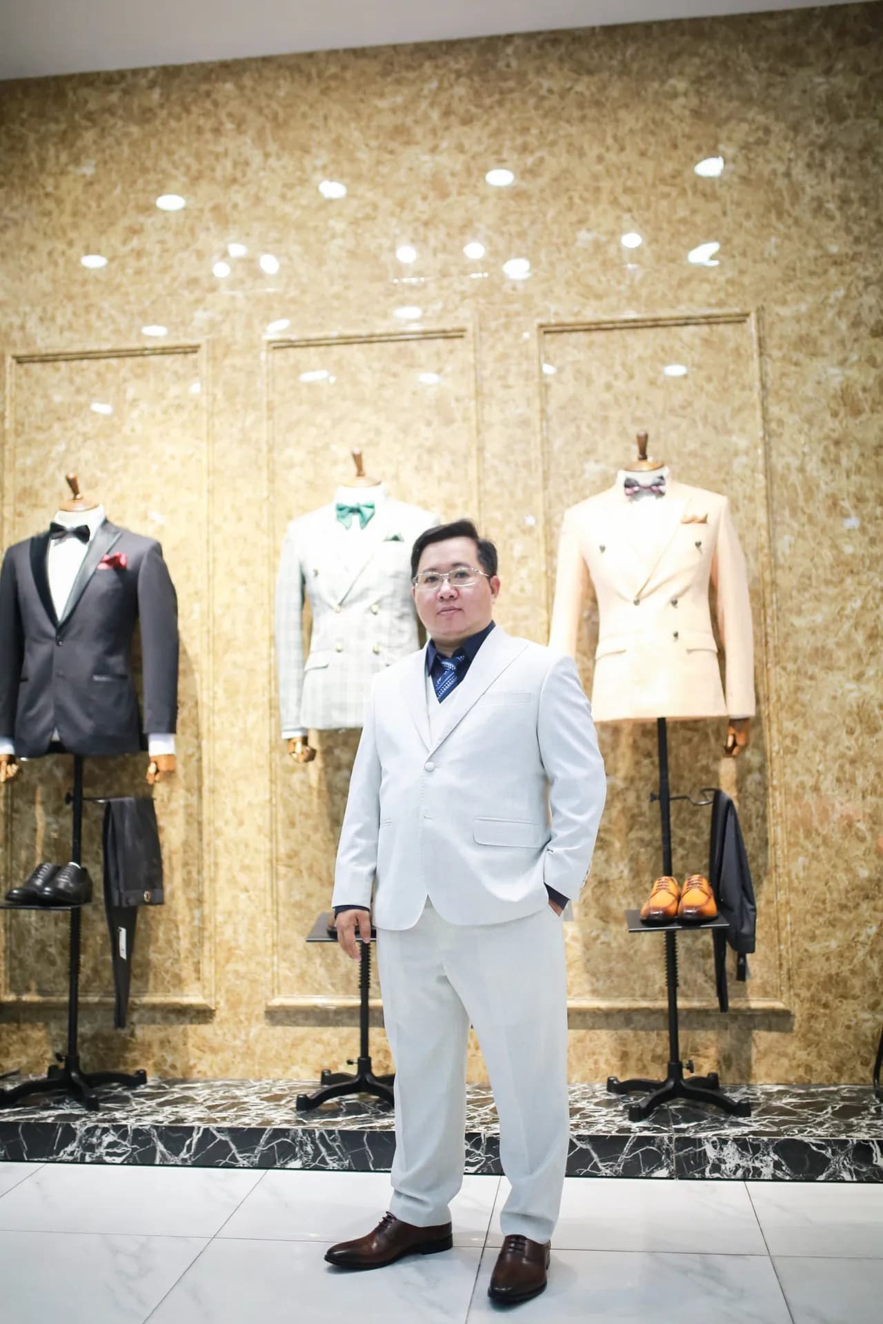 Suit trắng ba mảnh, cà vạt xanh, đứng giữa các manocanh tuxedo và vest hai hàng