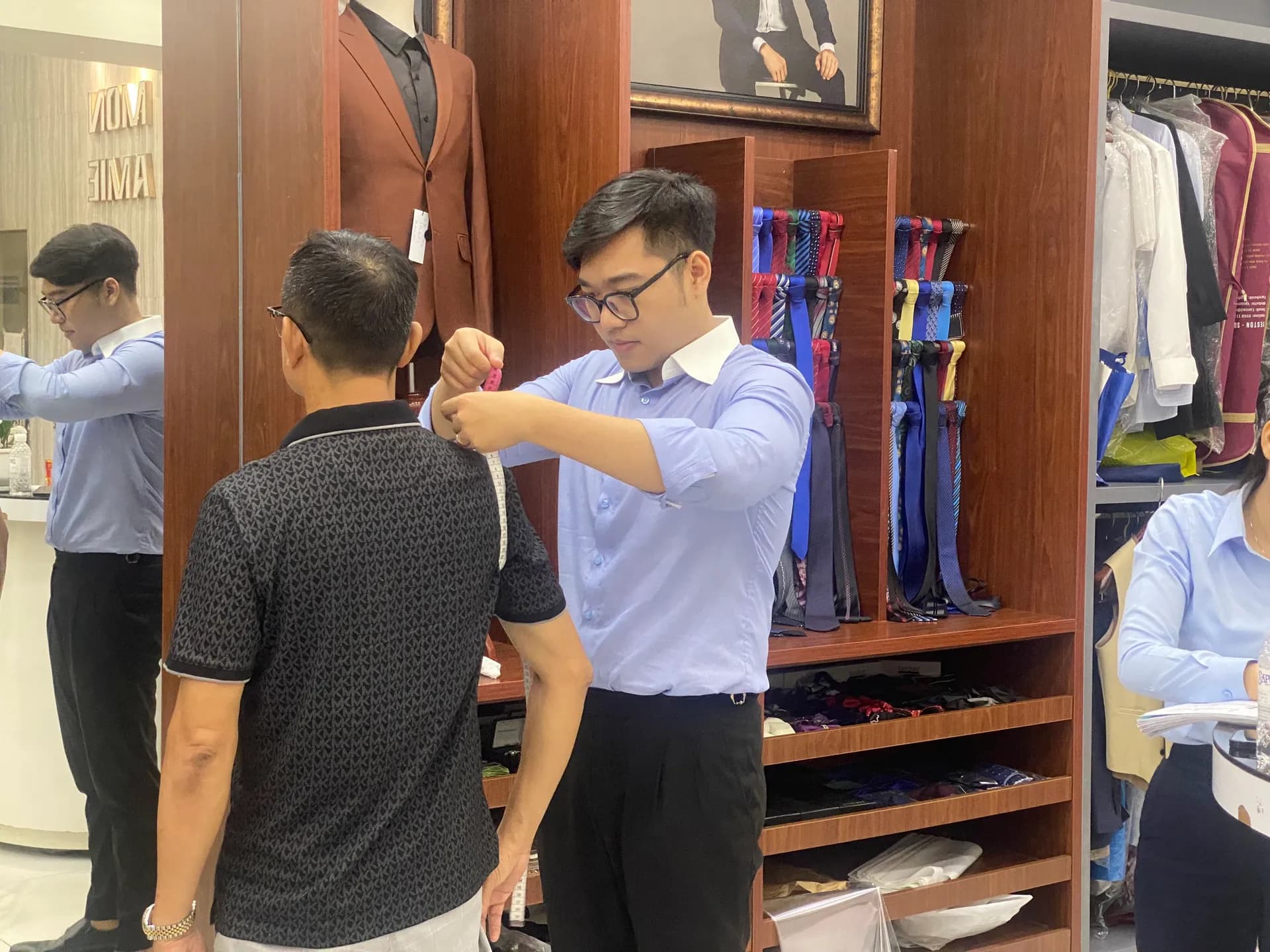 Nhân viên đo ngang vai cho khách tại showroom, áo sơ mi xanh nhạt