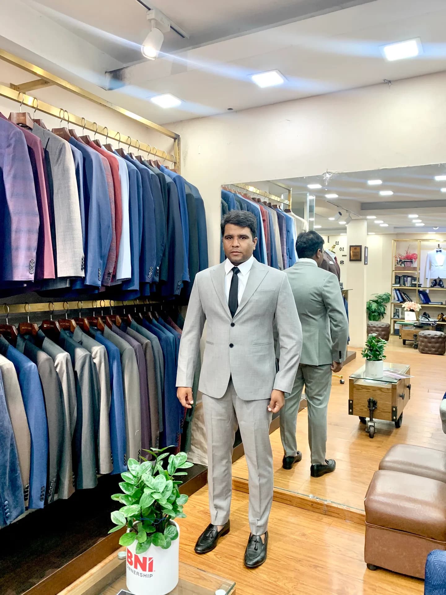 Suit xám nhạt hai khuy, cà vạt đen, đứng cạnh dãy vest treo