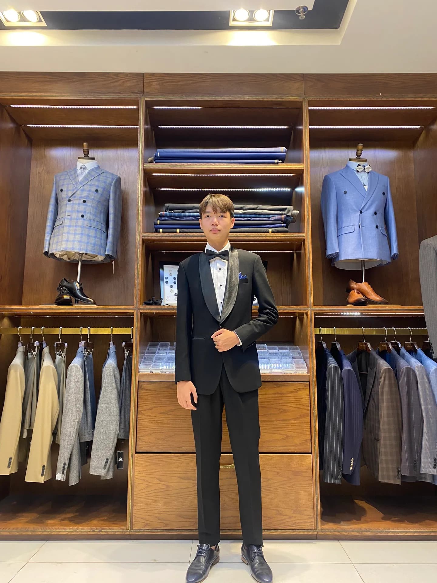 Tuxedo đen cổ lụa nơ đen, đứng trước kệ gỗ trưng bày vest caro xanh