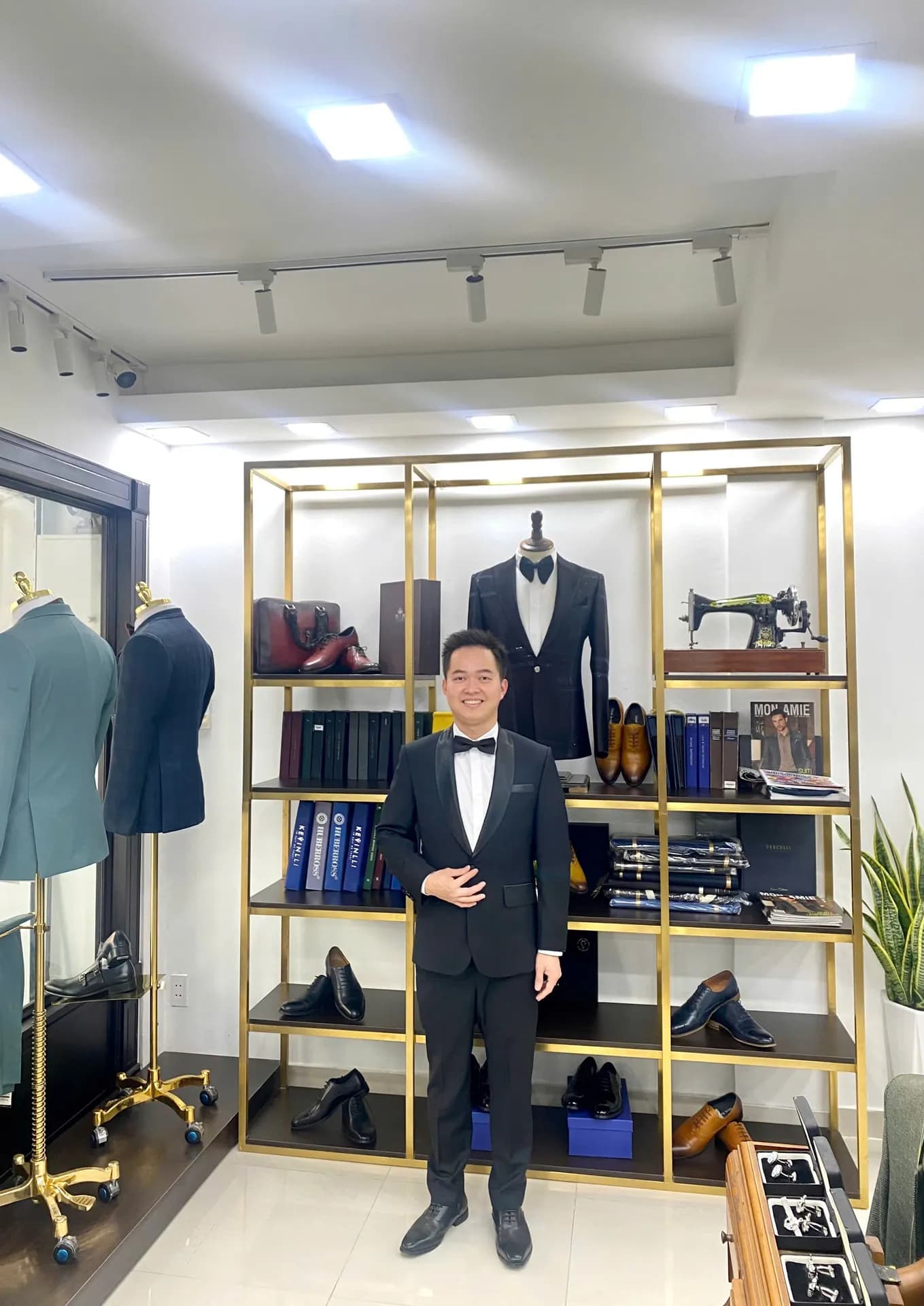 Tuxedo đen nơ đen, đứng trước kệ vàng đồng giày và vải trong showroom sáng