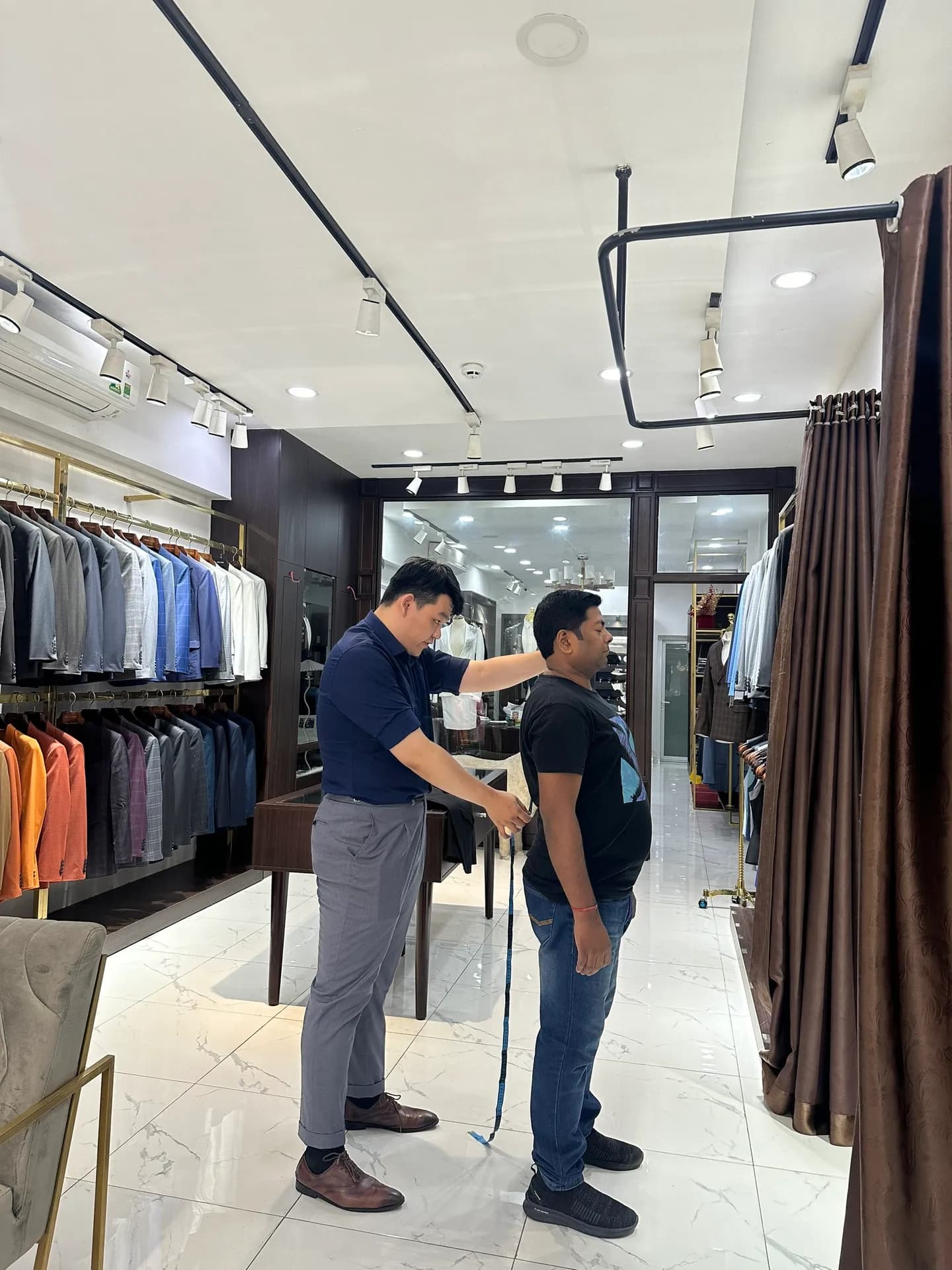 Nhân viên đo chiều dài thân cho khách trong showroom sáng, tủ vest hai bên