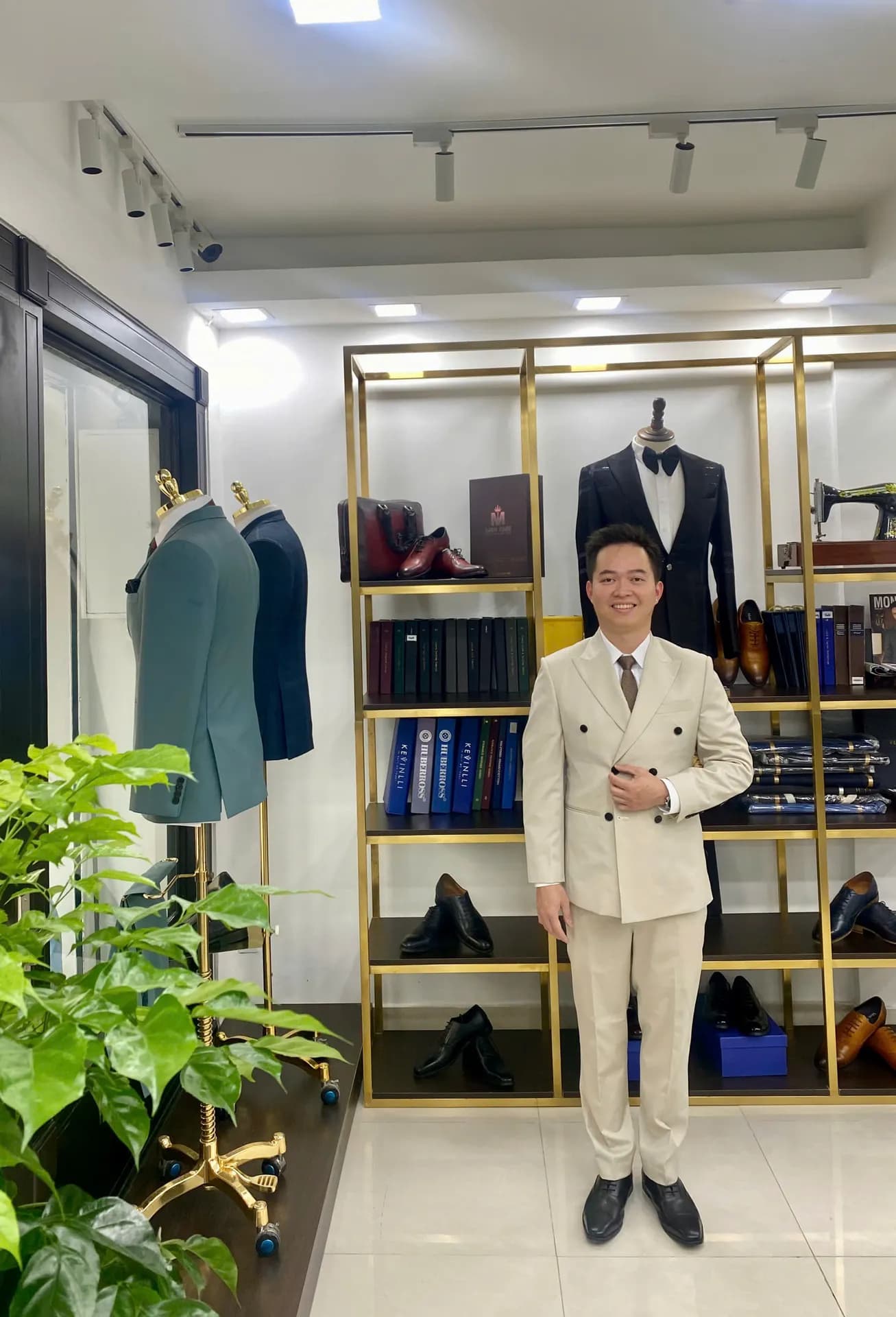 Suit kem hai hàng khuy, cà vạt nâu, đứng trước kệ vàng đồng