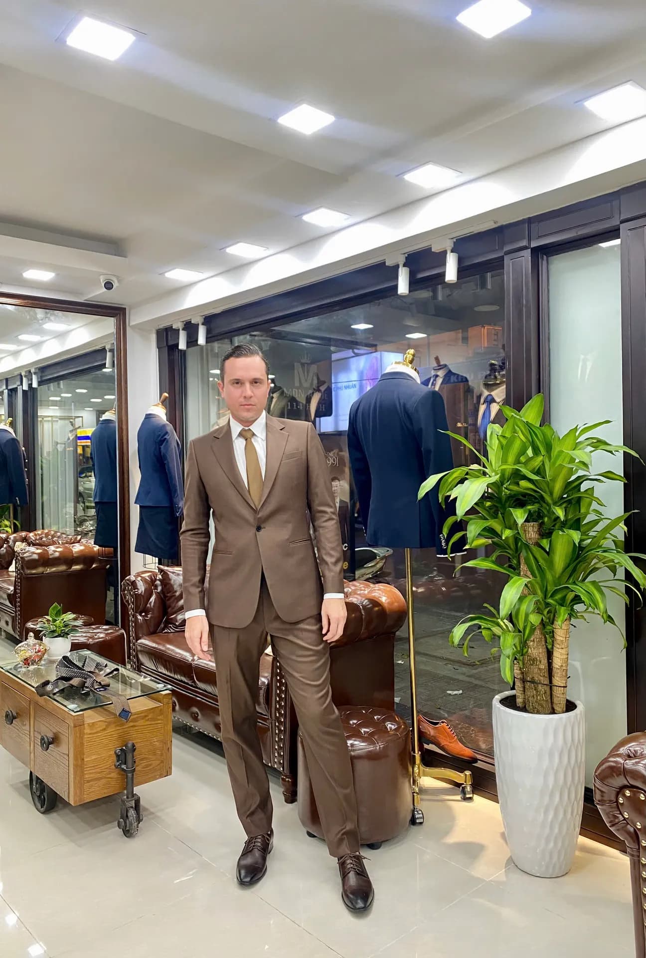 Khách mặc suit nâu cà vạt vàng đồng giữa showroom, nền sofa da