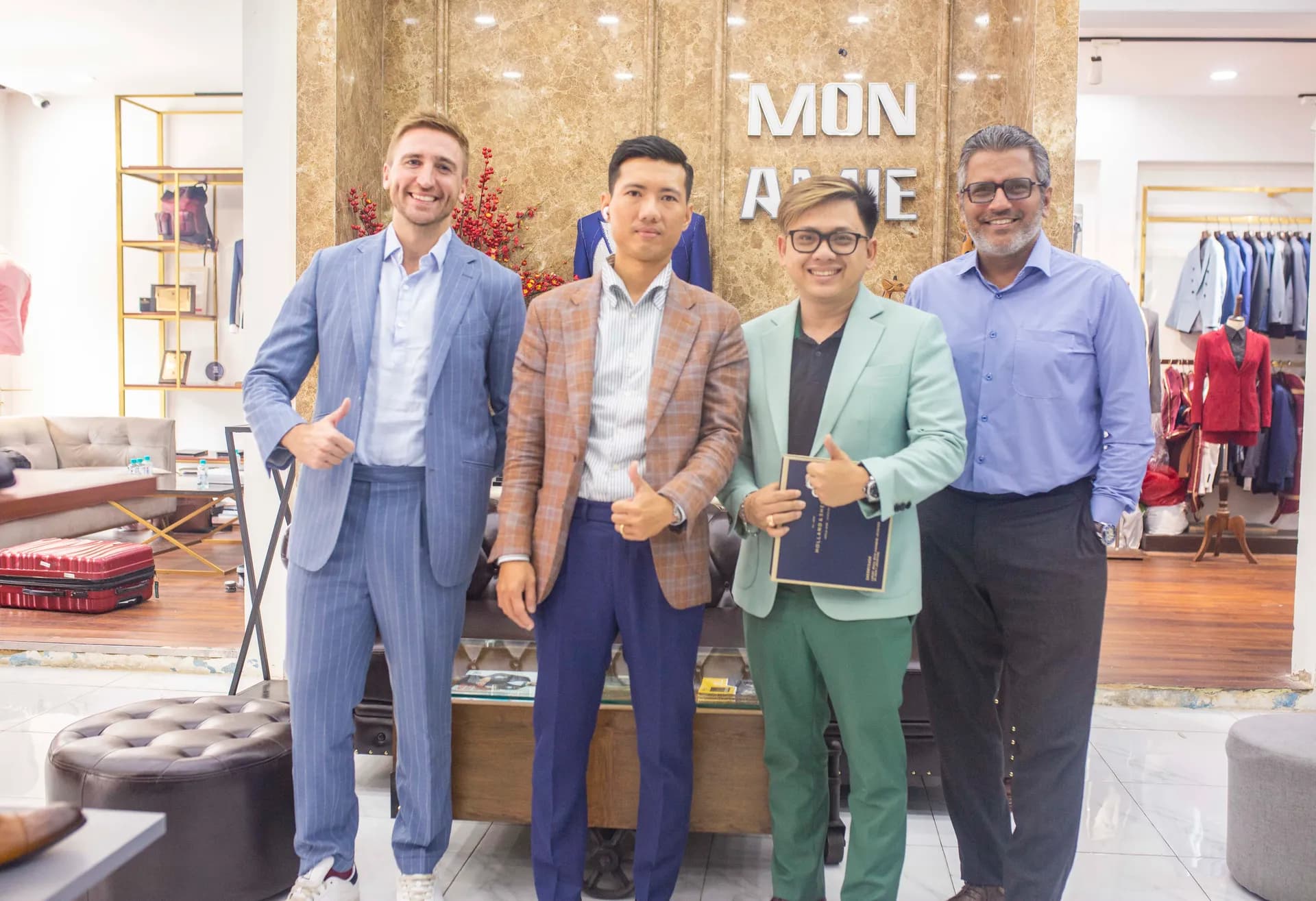 Bốn khách mặc suit nhiều sắc màu giơ ngón cái trước biển Mon Amie