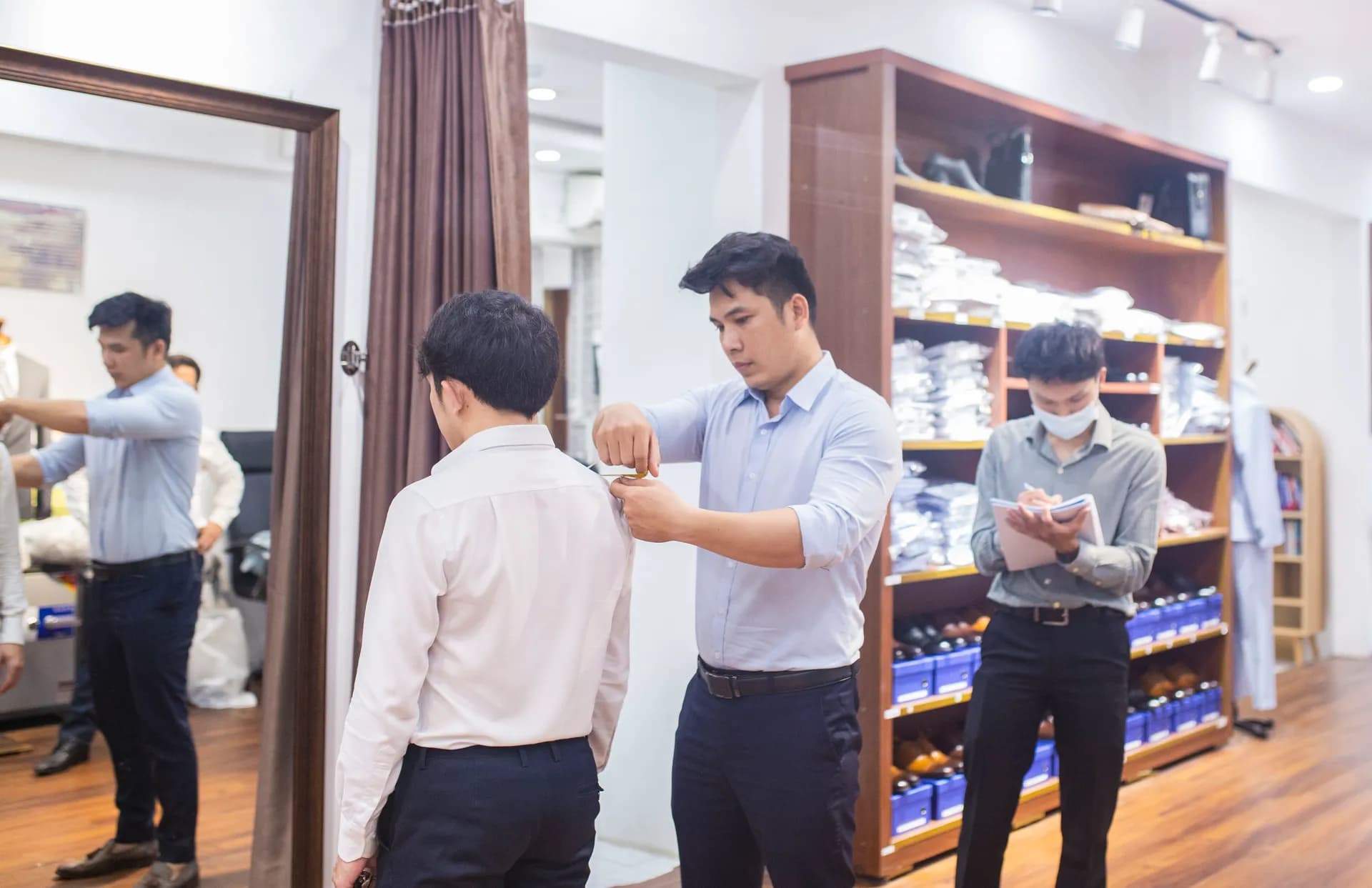 Nhân viên đo vai cho khách sơ mi trắng, người ghi số đo bên cạnh