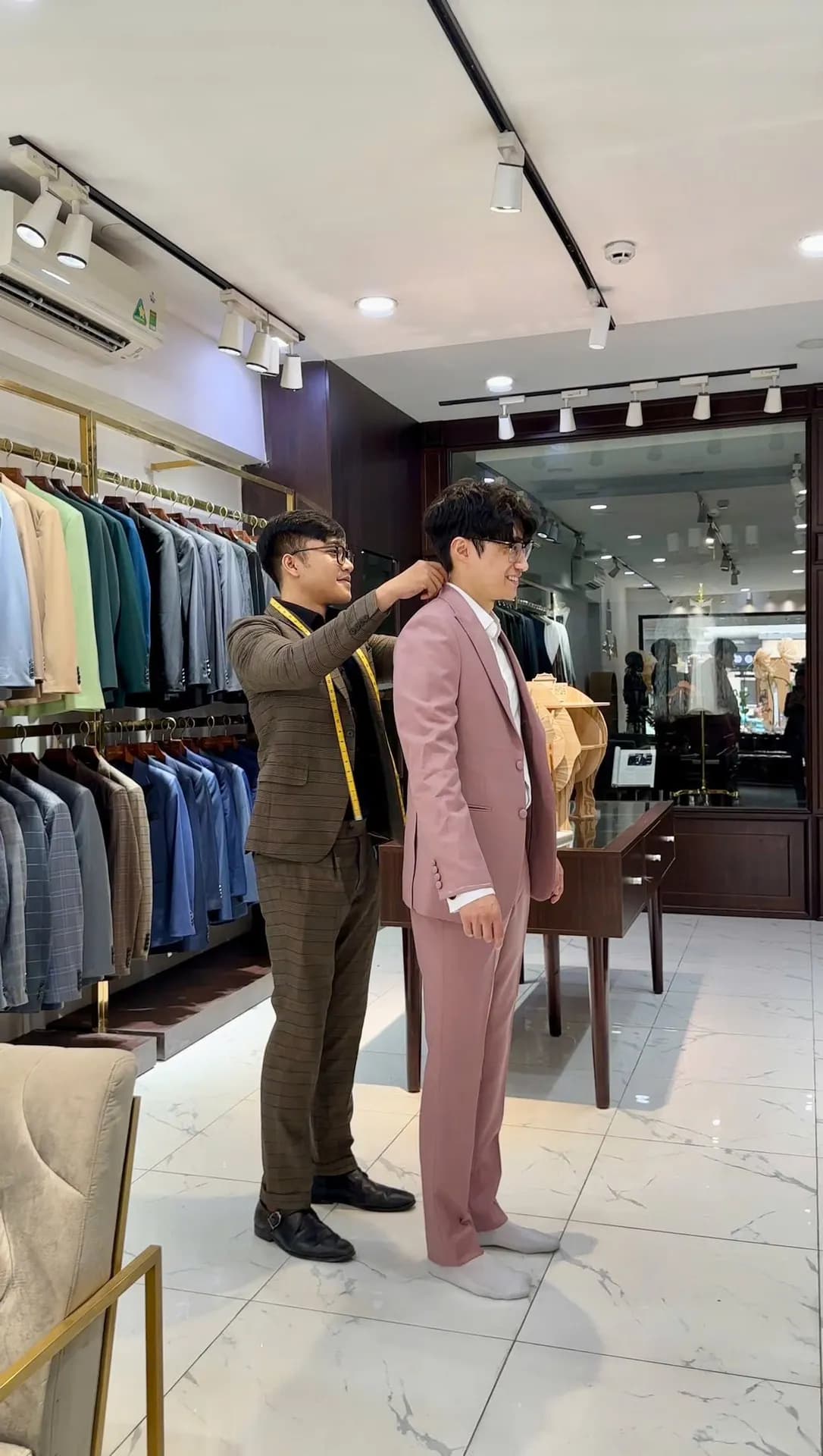 Thợ may đeo thước dây chỉnh vai suit hồng cho khách thử đồ