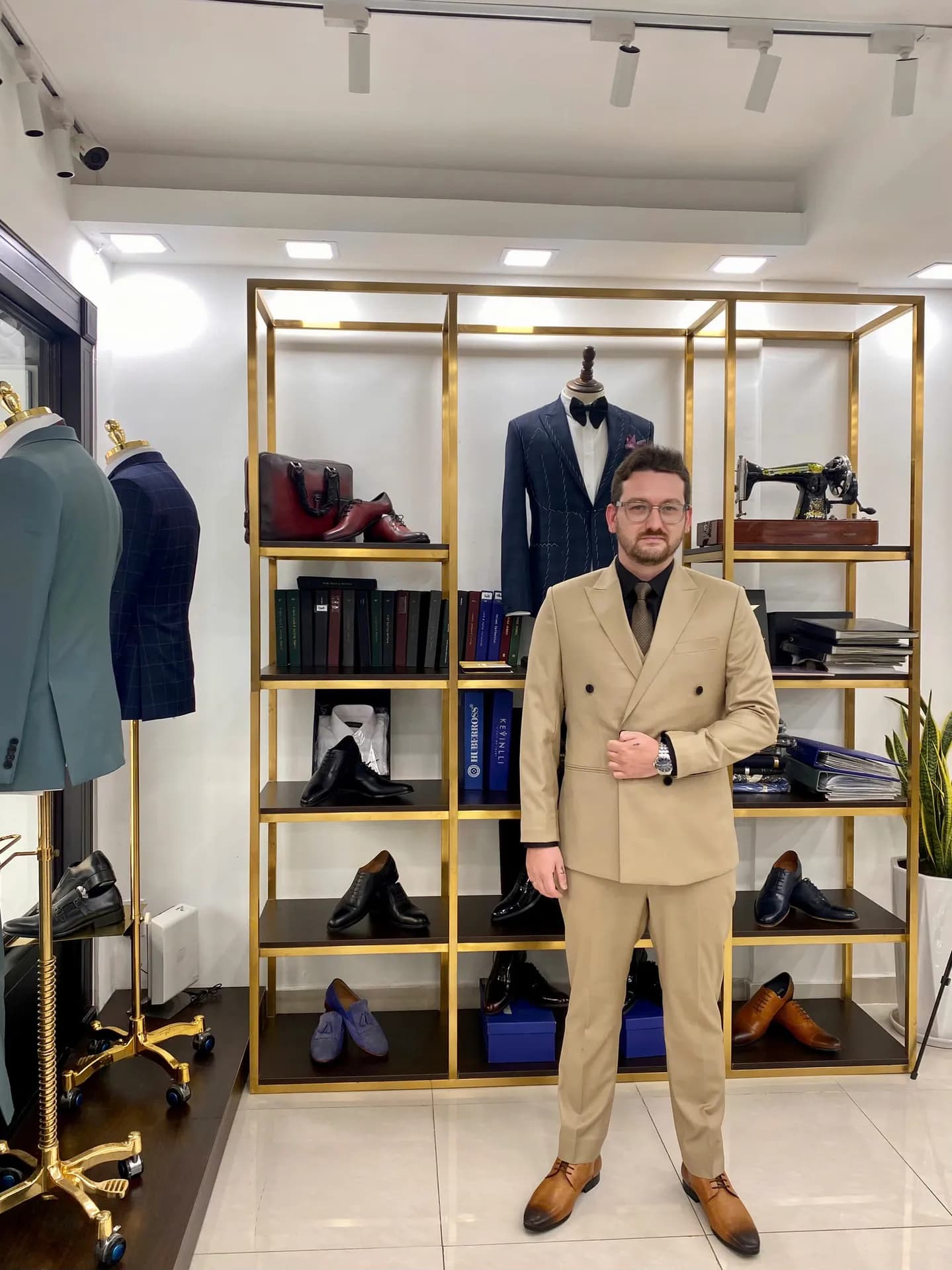 Khách mặc suit be hai hàng, sơ mi đen, đứng cạnh kệ vàng đồng