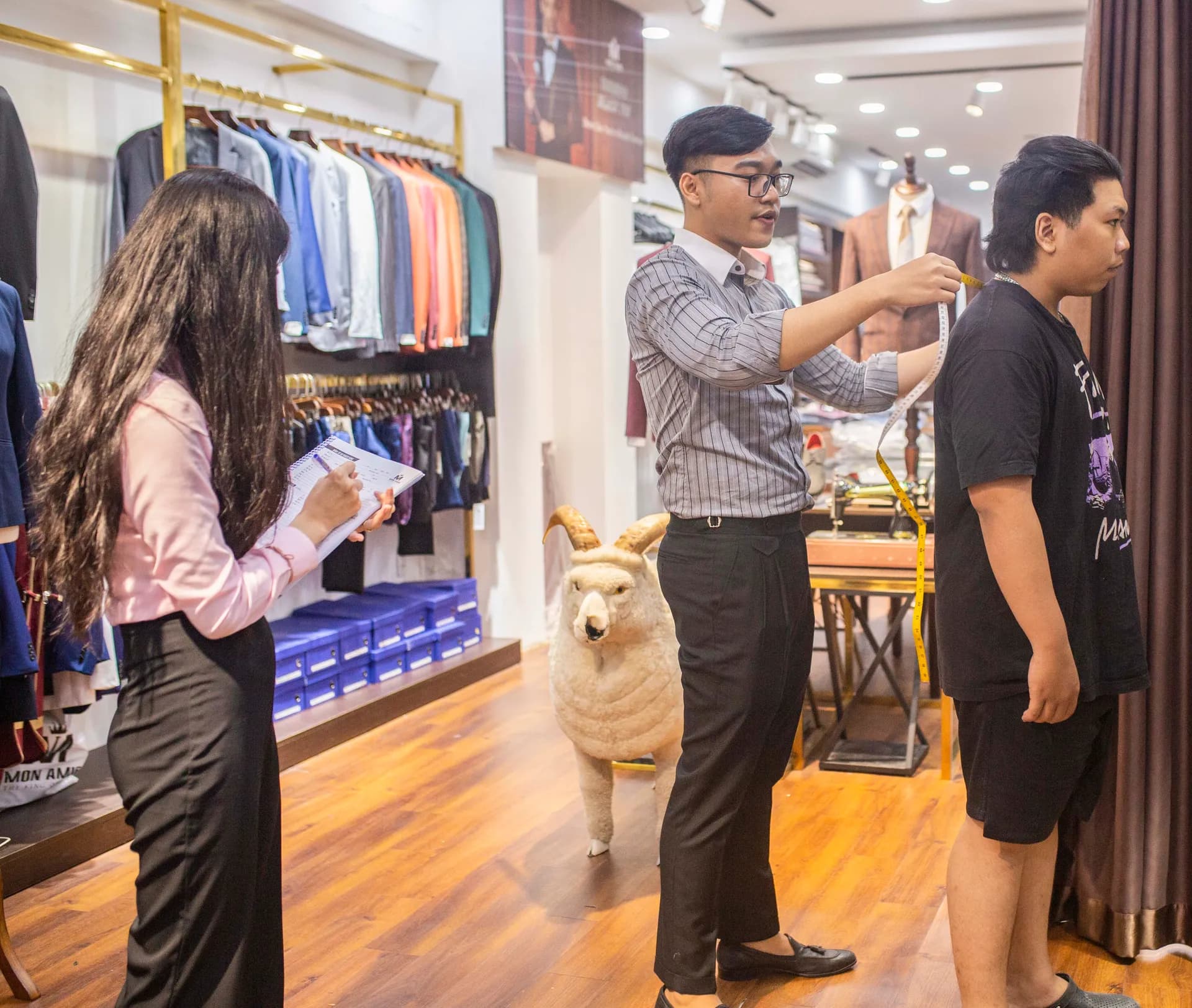 Thợ may đo vai cho khách, nhân viên ghi số đo bên cạnh
