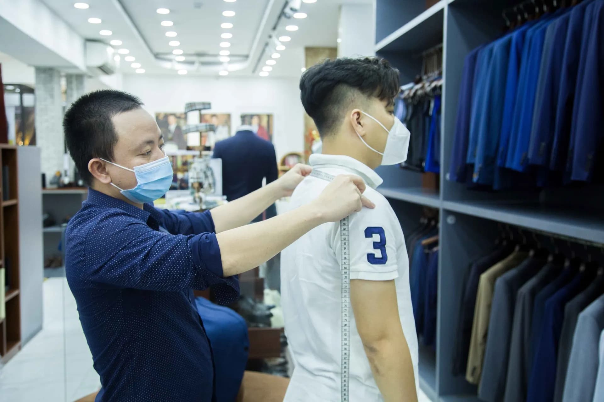 Nhân viên đo vai cho khách, kệ vest xanh navy phía sau