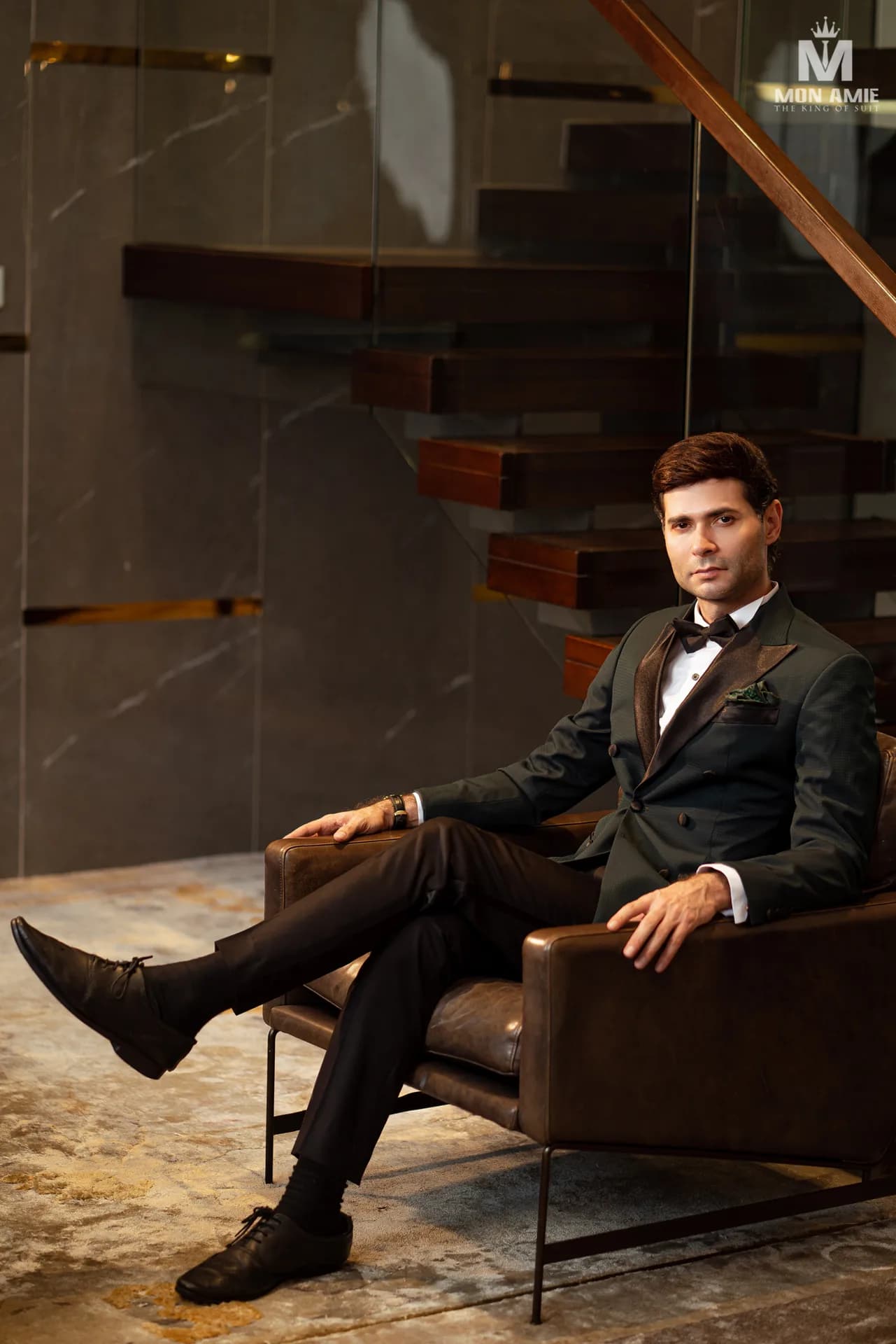 Tuxedo xanh rêu hai hàng khuy, ve satin, nơ đen, ngồi ghế da sang trọng.