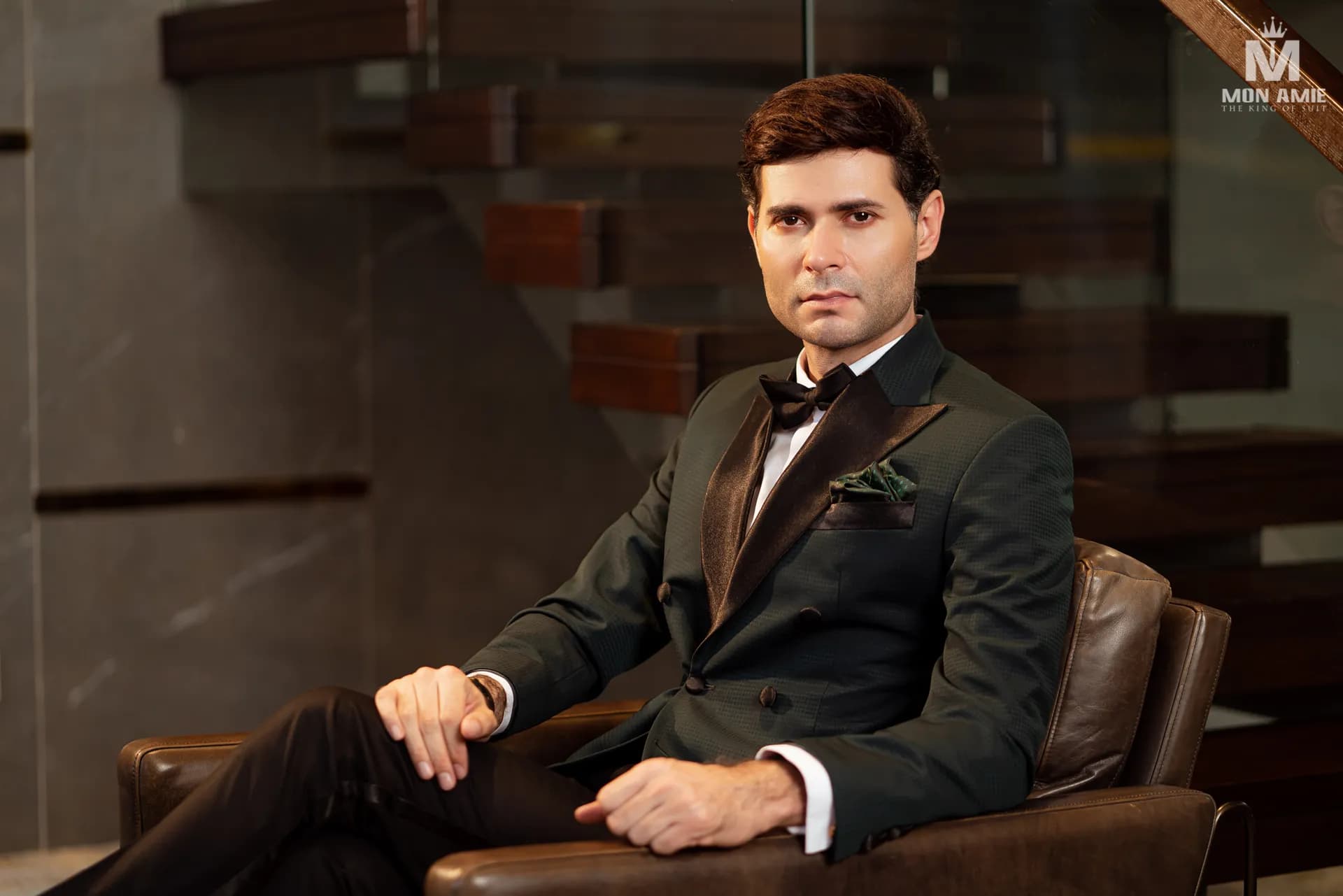 Tuxedo xanh rêu ve nhung, nơ đen, ngồi ghế da bên cầu thang gỗ