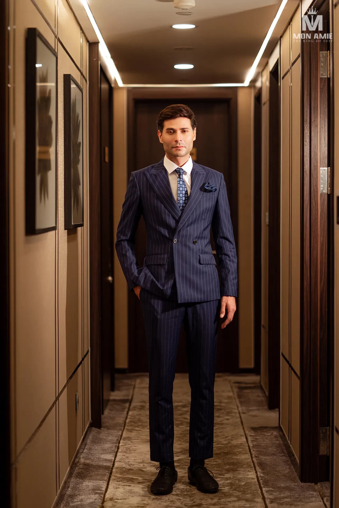 Suit hai hàng khuy xanh navy đứng giữa hành lang khách sạn ánh vàng