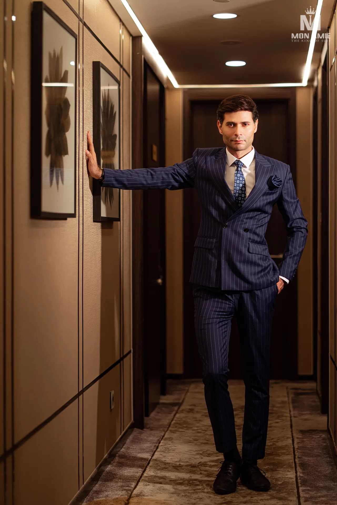 Suit xanh navy ba mảnh, một tay chống tường trong hành lang gỗ