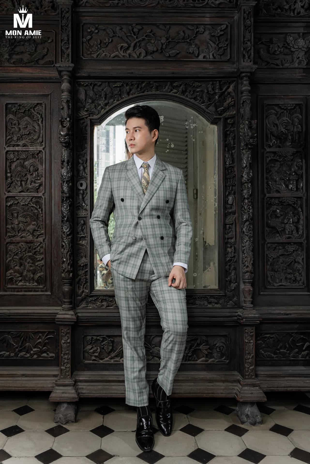 Vest caro xanh lá nhạt toàn thân trước tủ gỗ chạm, sàn gạch hoa
