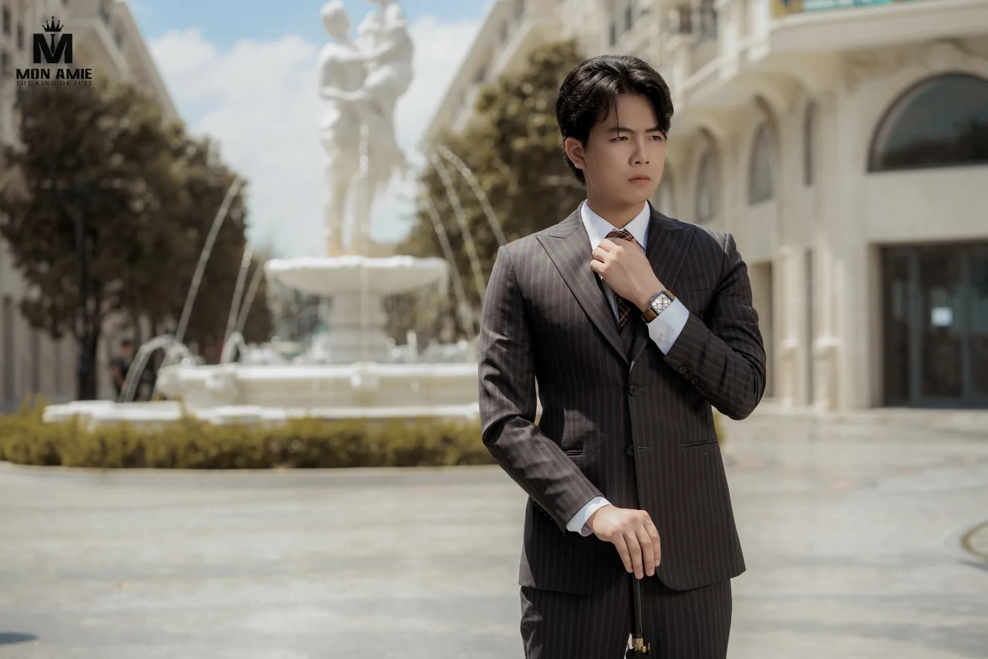 Suit sọc nâu đậm, chỉnh cà vạt, đài phun nước trắng phía sau
