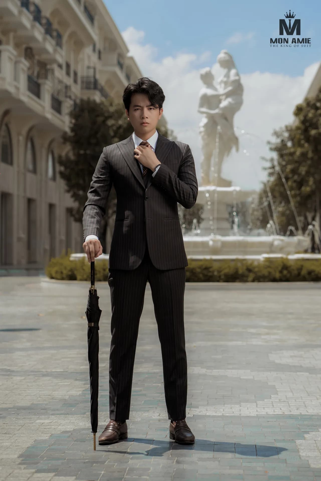 Suit nâu sọc ba mảnh, cầm ô đen, đứng bên tượng đài phun nước