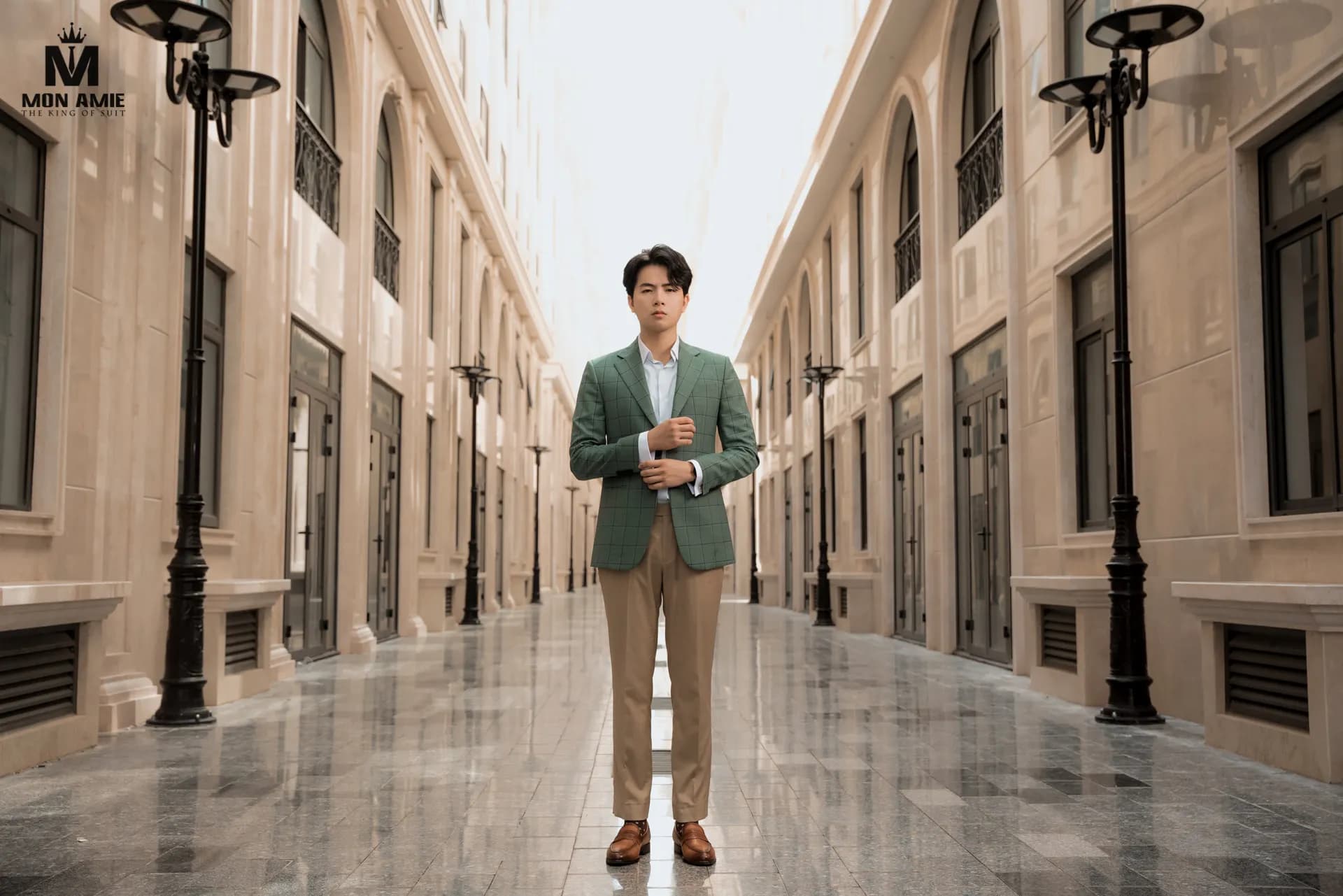 Blazer xanh lá, quần kaki, đứng giữa dãy phố vòm, khung ngang
