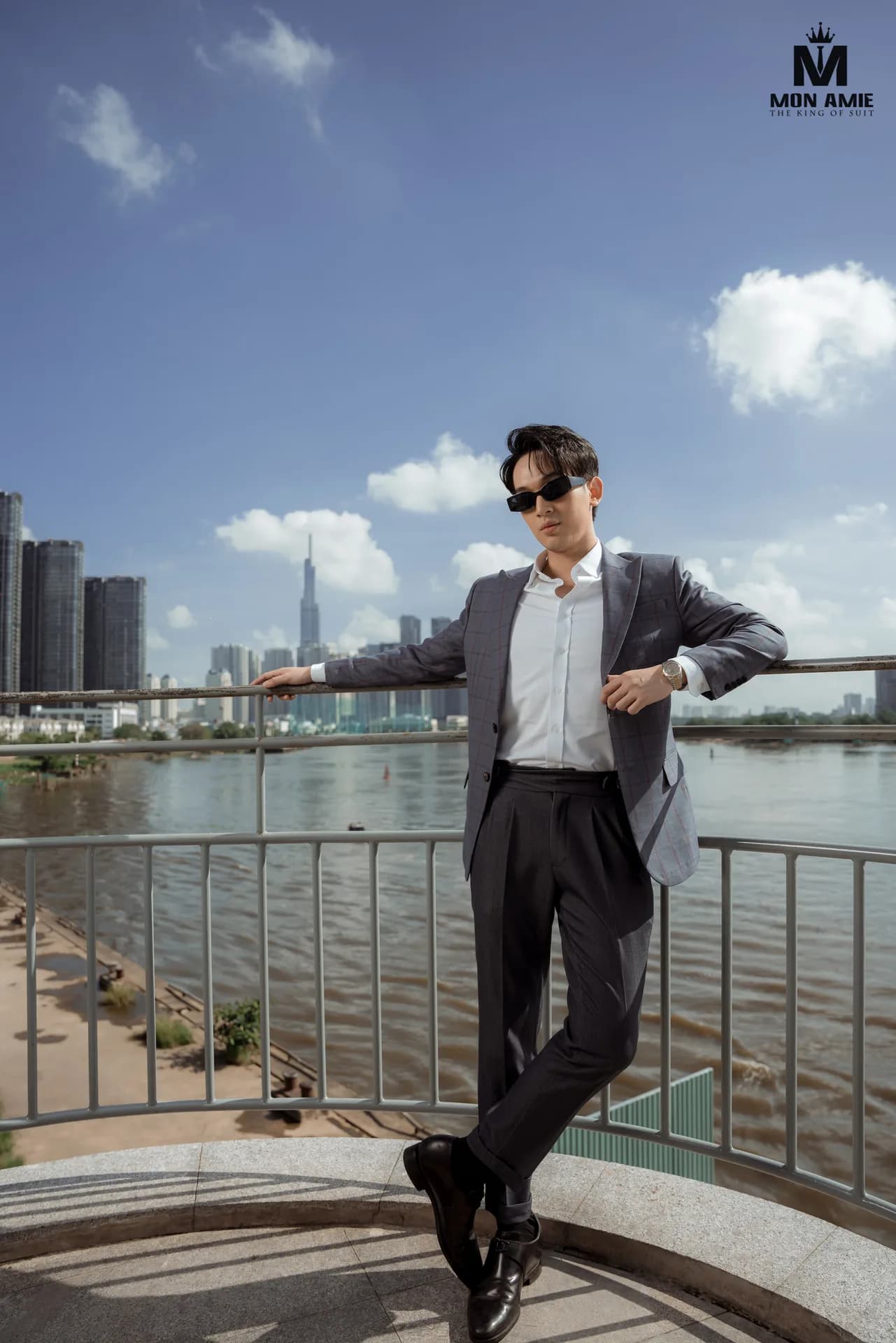 Blazer xám kẻ ô, quần ống đứng, tựa lan can nhìn sông và đường chân trời thành phố